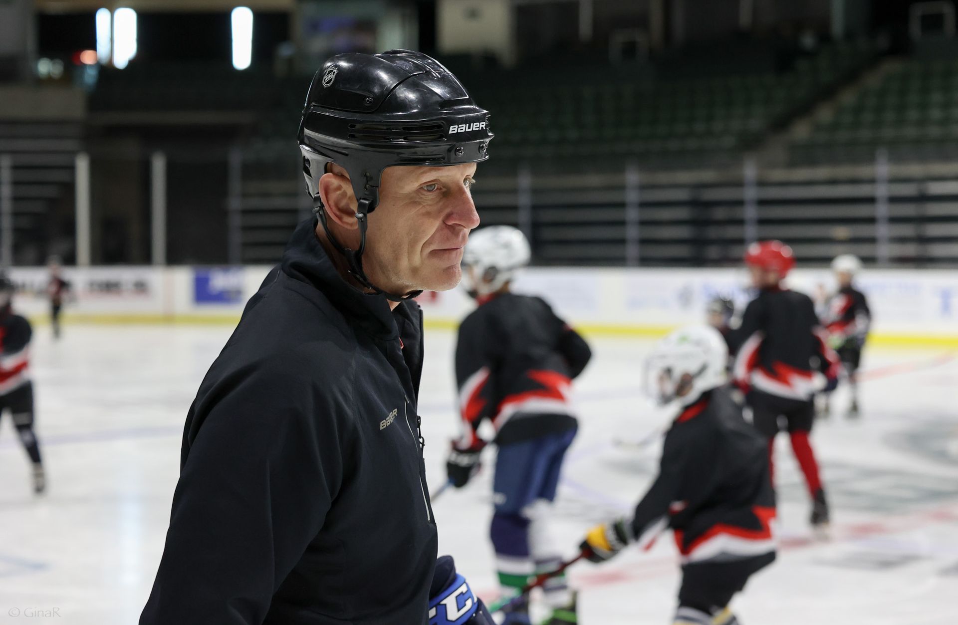 Hockey coach in black helmet and jacket stands on the ice while players practice in the background