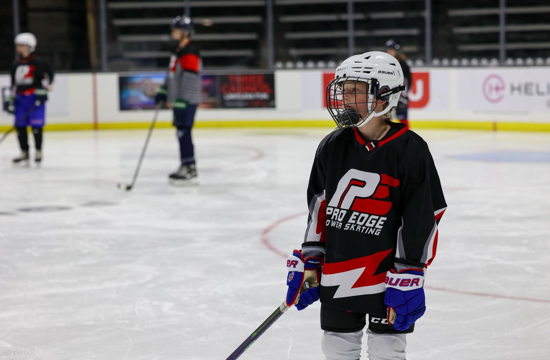 Child in black hockey jersey and helmet standing on an ice rink with a stick, other players blurred in background