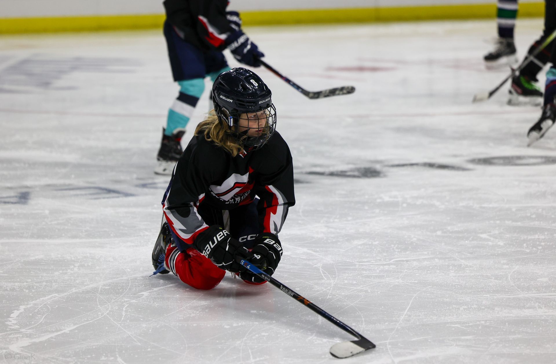 Youth hockey player in black and red kneels on ice, holding a stick during a game.