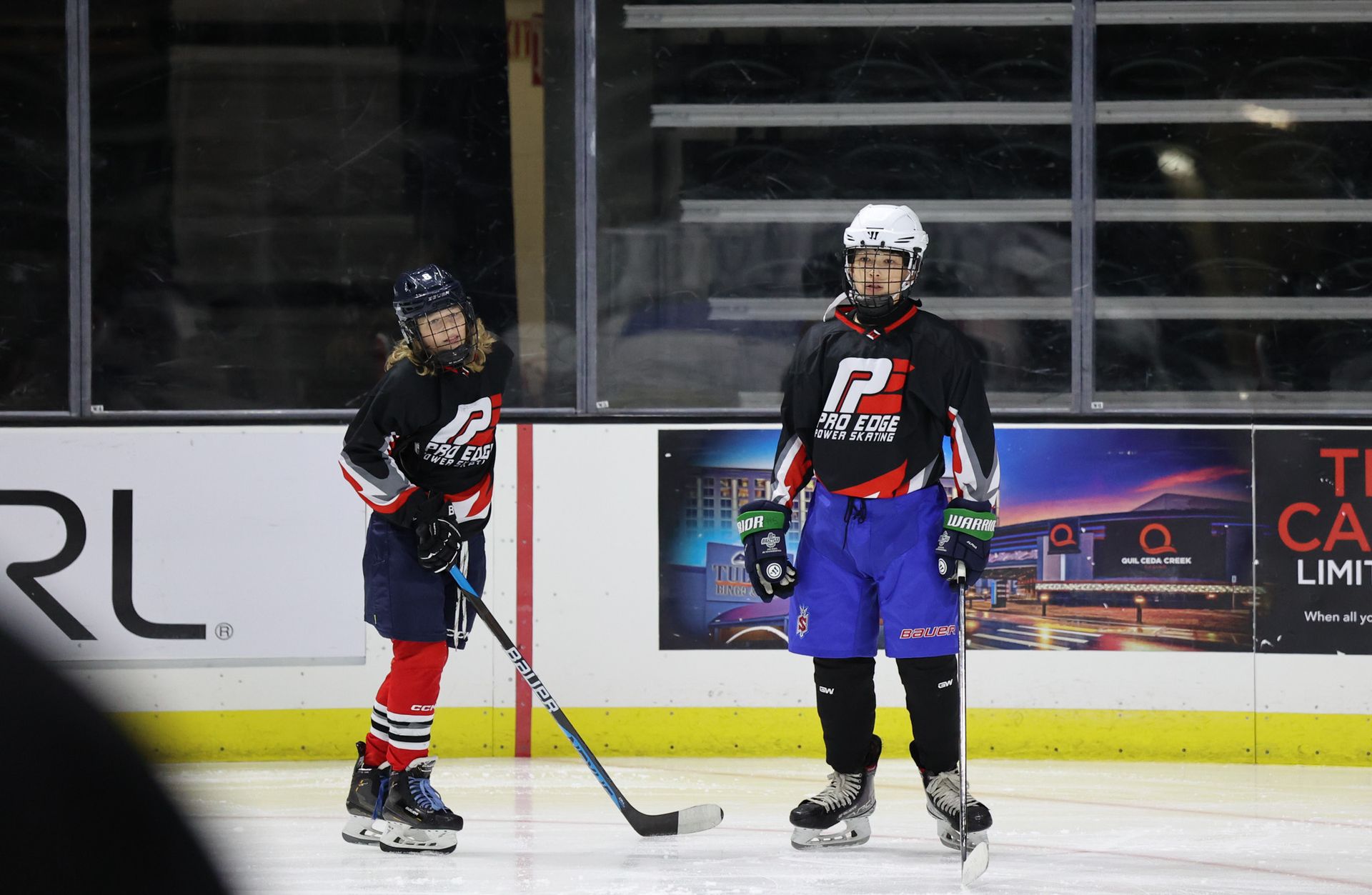 Two hockey players in black jerseys stand on an indoor rink with sticks and helmets, facing each other.