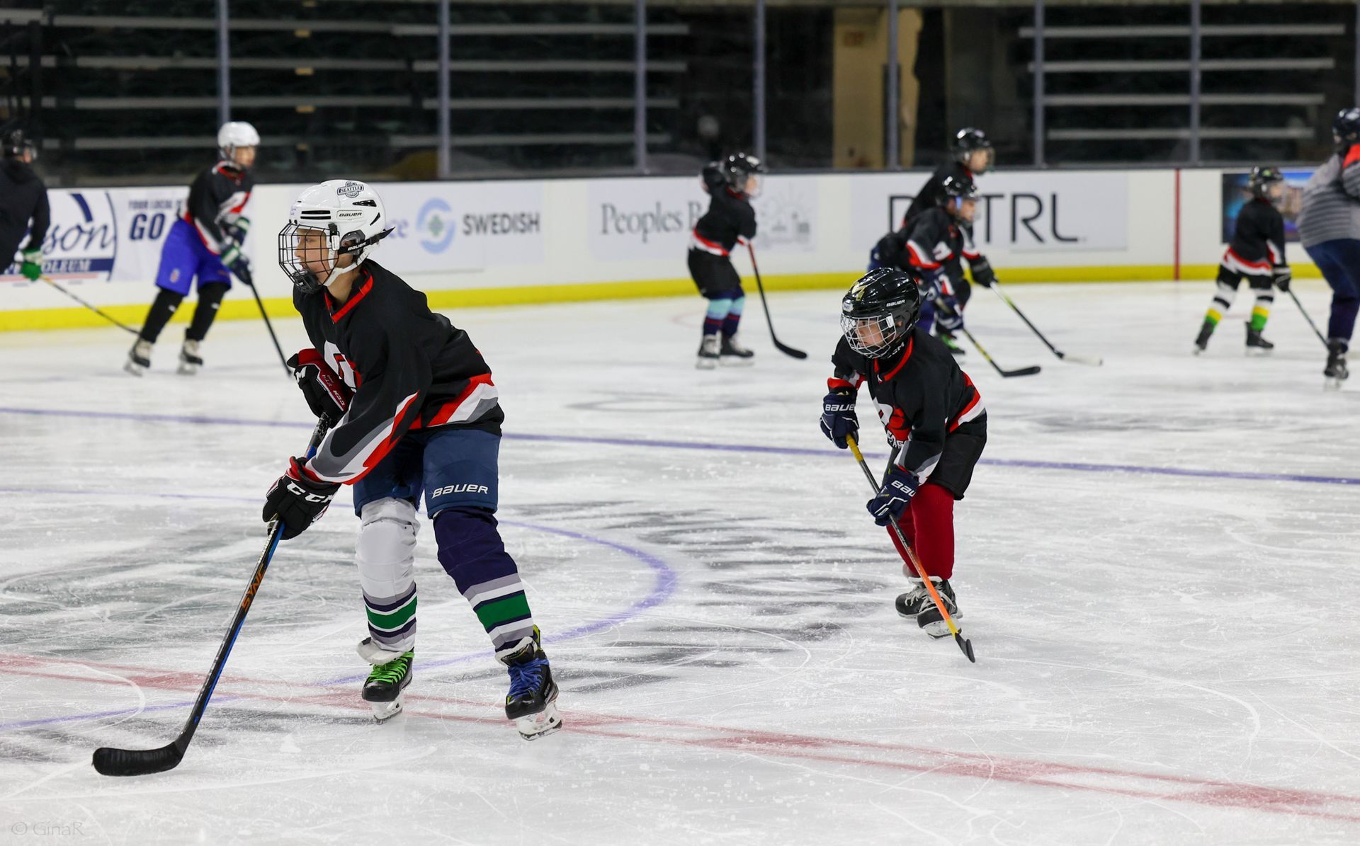 Hockey players in black and red skate during a game on an indoor ice rink.