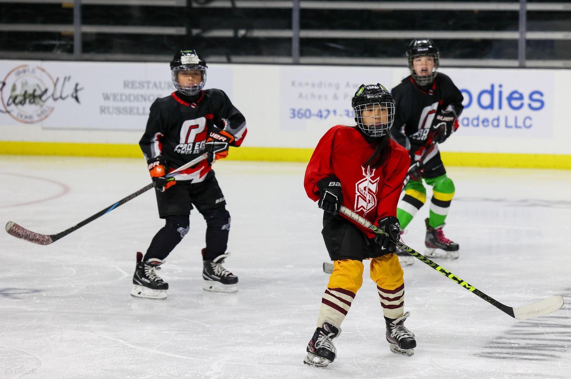 Three youth hockey players skating on ice, one in red centered with two in black jerseys behind.