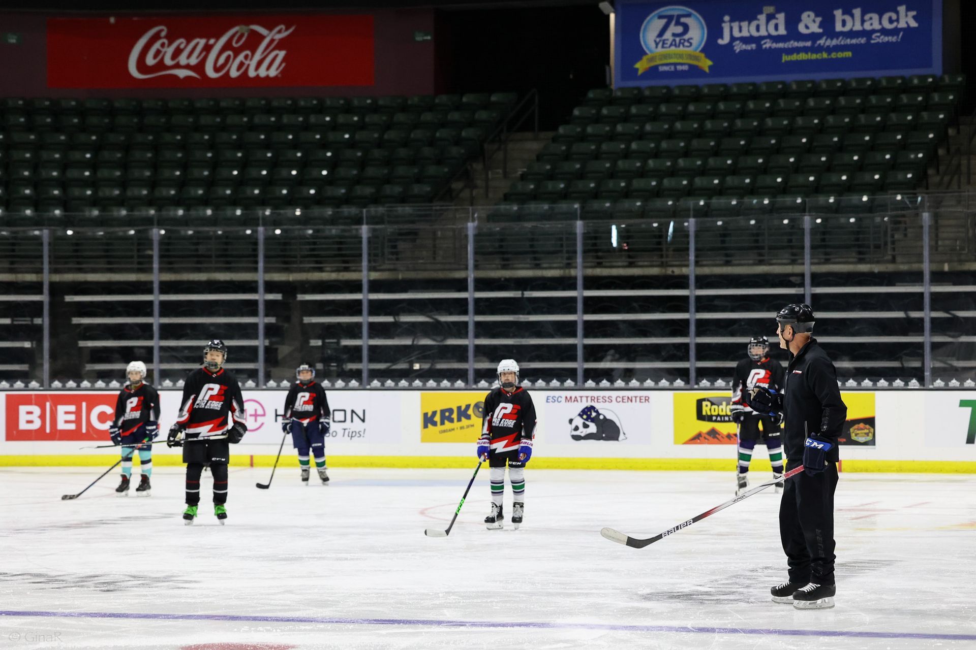 Ice hockey players and a coach stand on the rink during practice in an empty arena.
