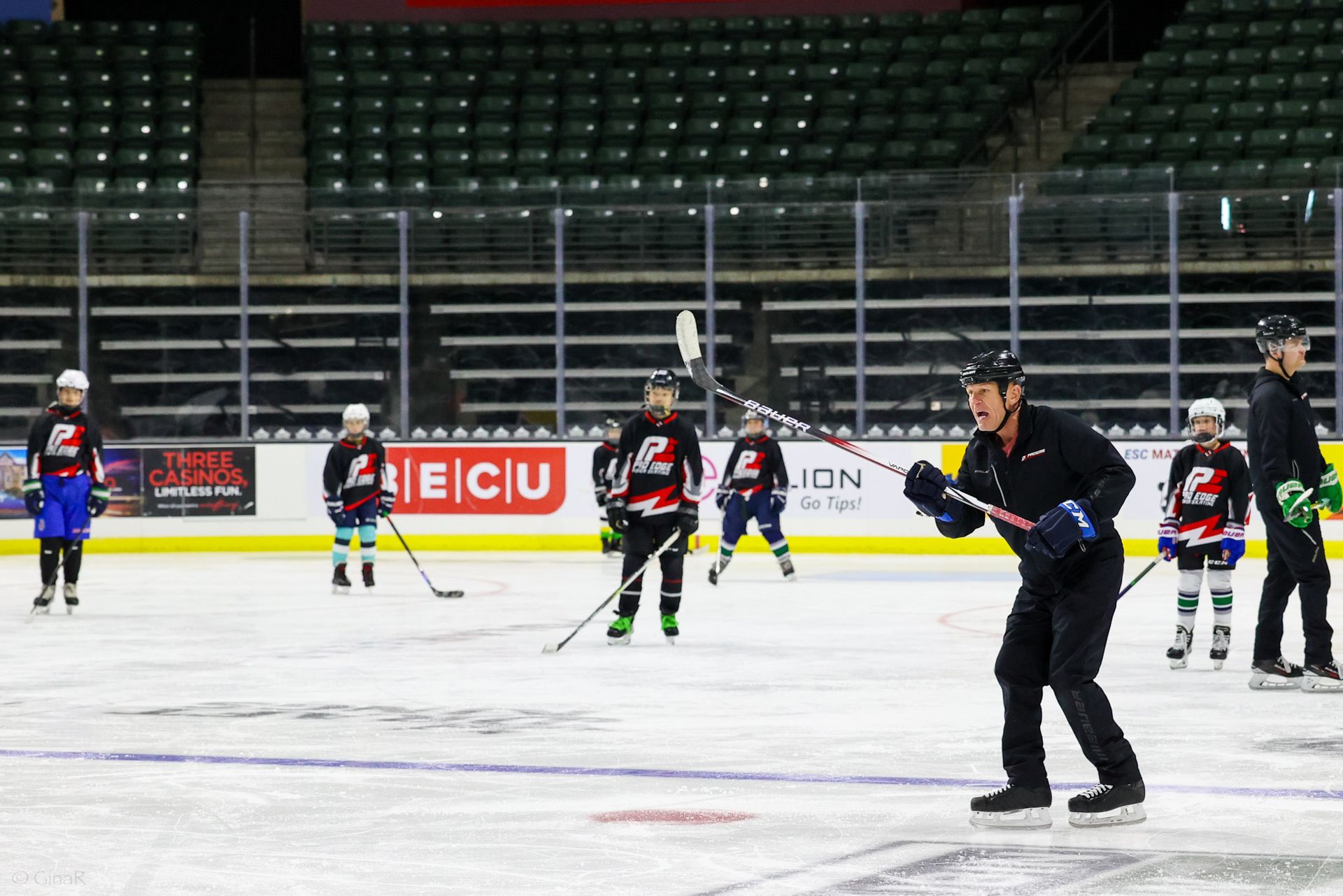 Ice hockey practice on an indoor rink, with players in black, red, and blue jerseys skating near the boards.