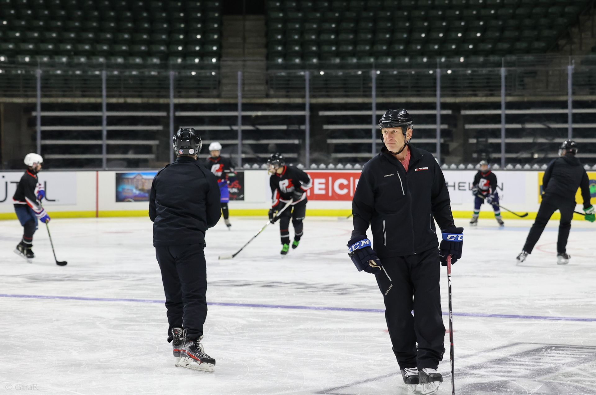 Ice rink practice with several skaters and coaches in black jackets on the ice