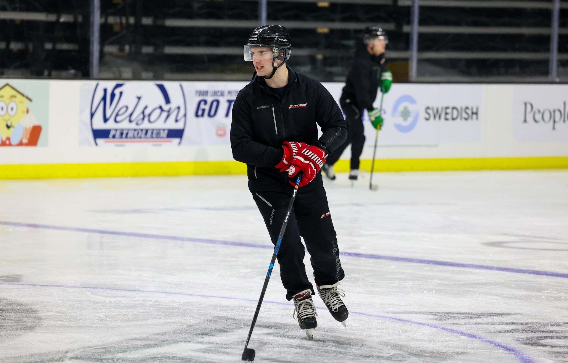 Ice hockey player in black gear skating on the rink with a red glove, holding a stick.