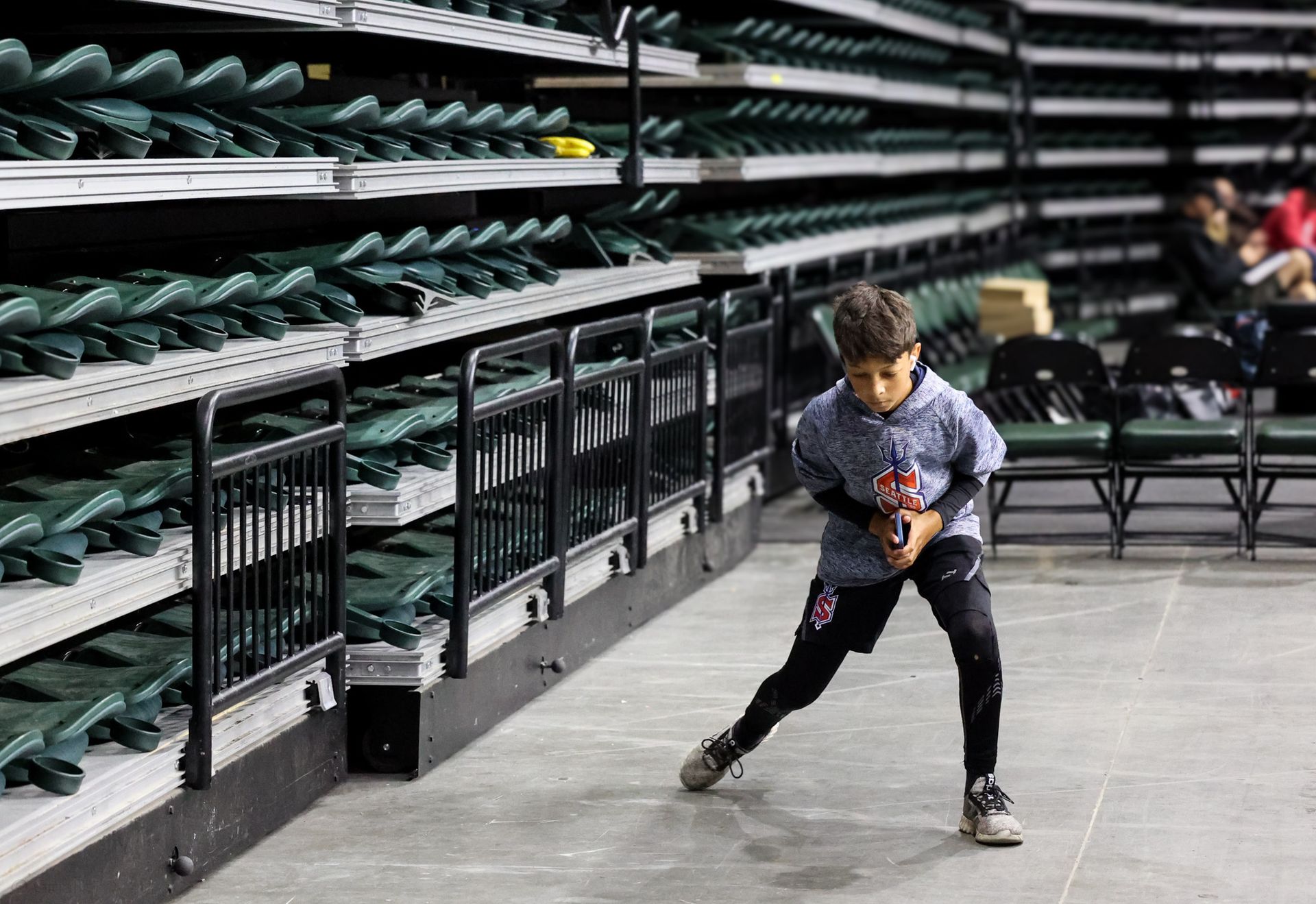 Young boy in a gray hoodie runs on a concrete walkway beside empty stadium seats.