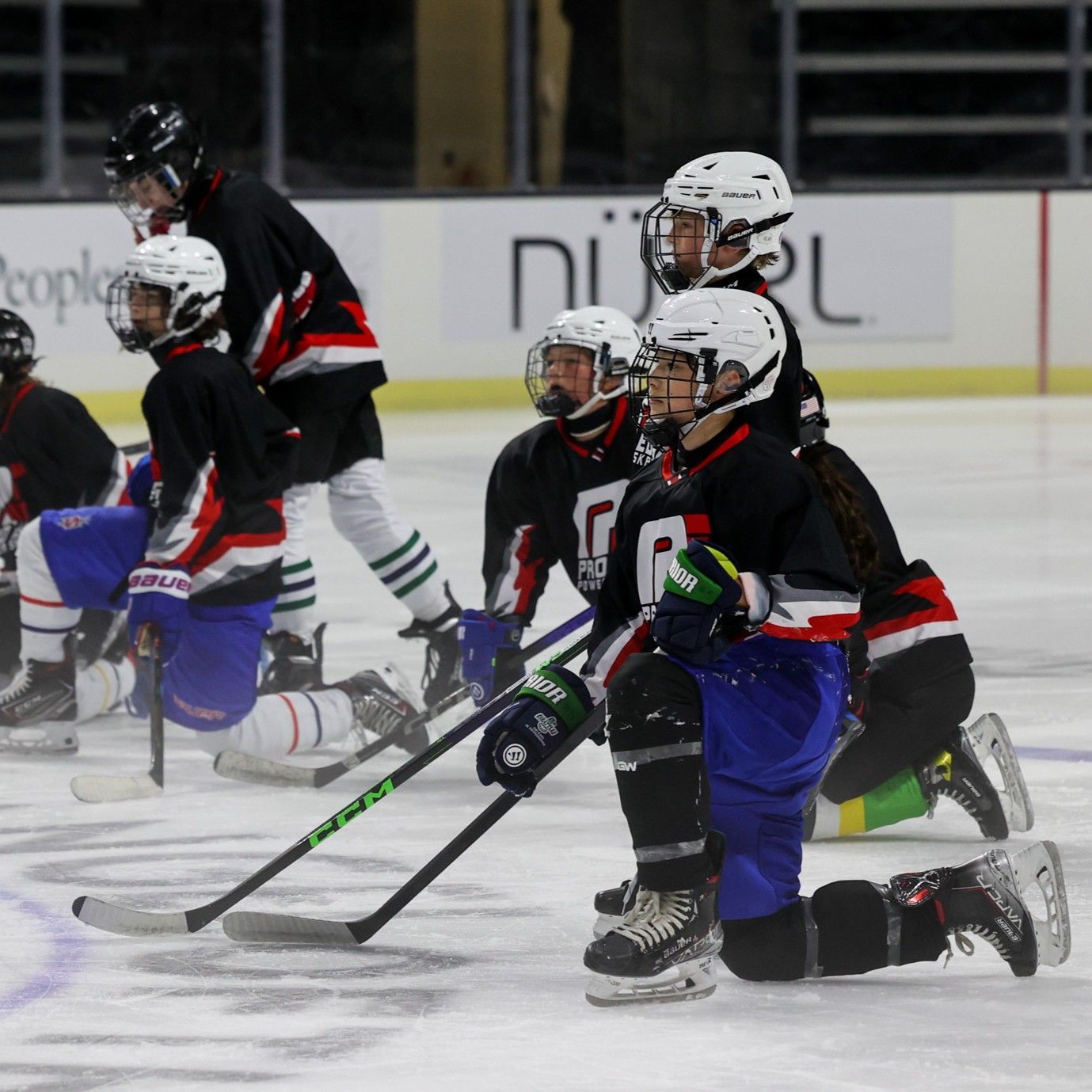 Inline hockey players in black and red jerseys crouch on an ice rink during play