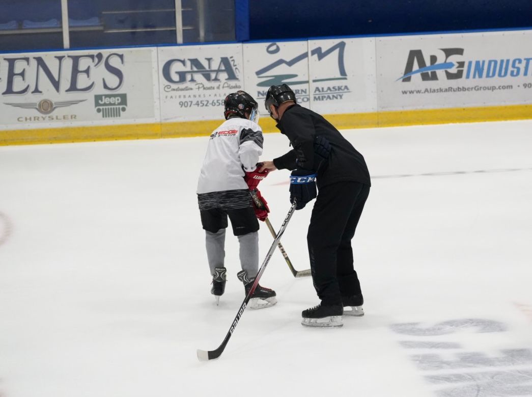 Two hockey players on an ice rink, one helping the other balance with a stick.