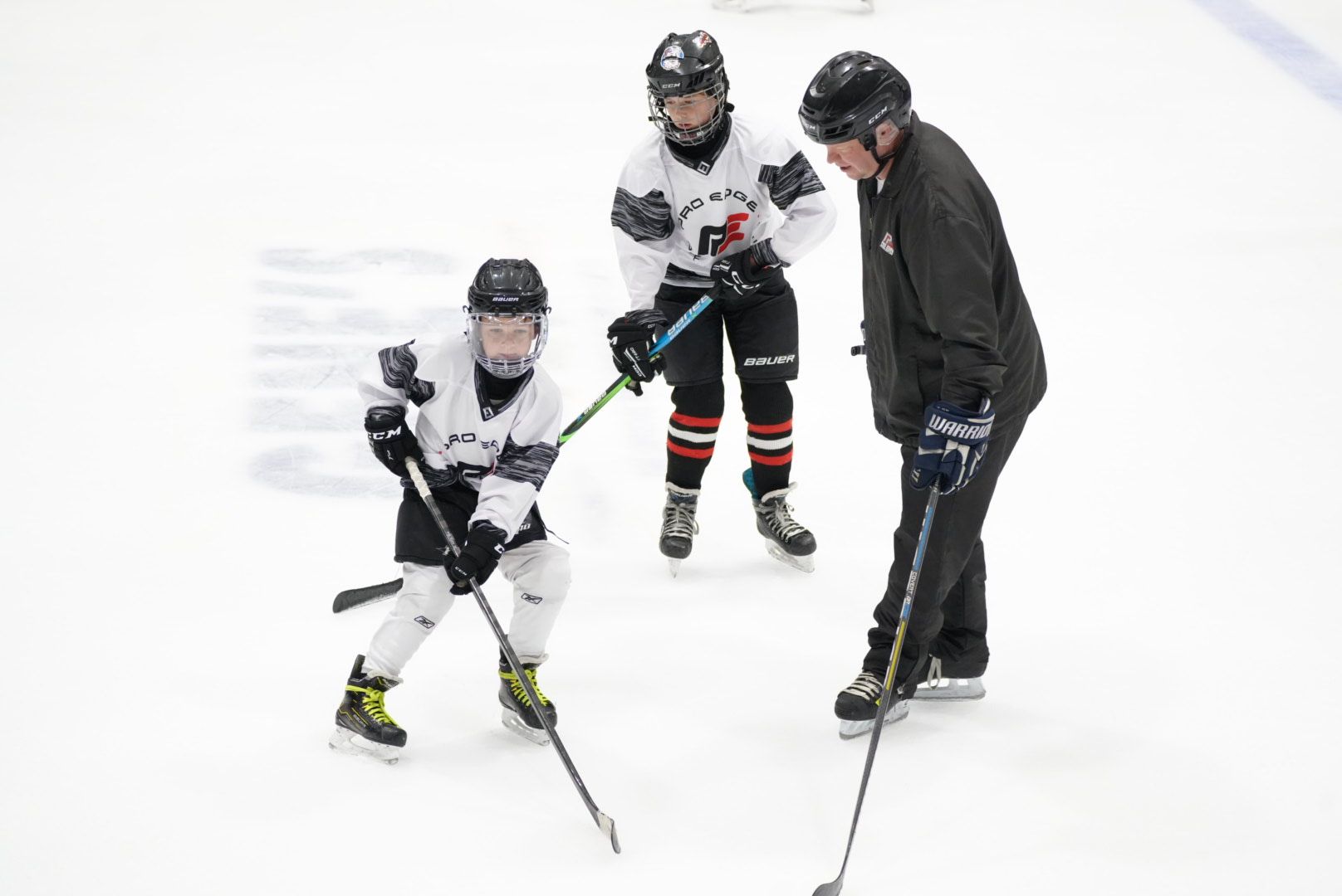 Three people on ice skates, including a coach, standing on an indoor ice rink.