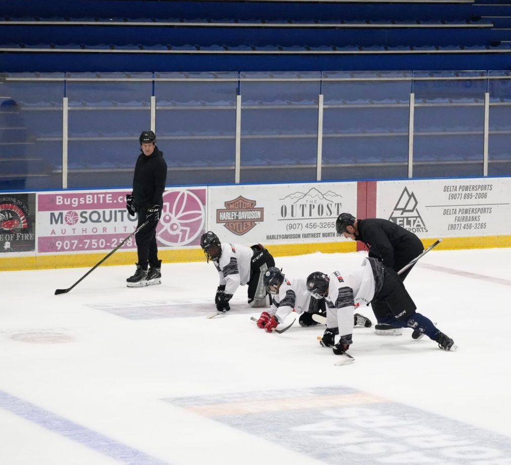 Youth hockey players crouch at the faceoff circle on an indoor ice rink while a coach watches nearby