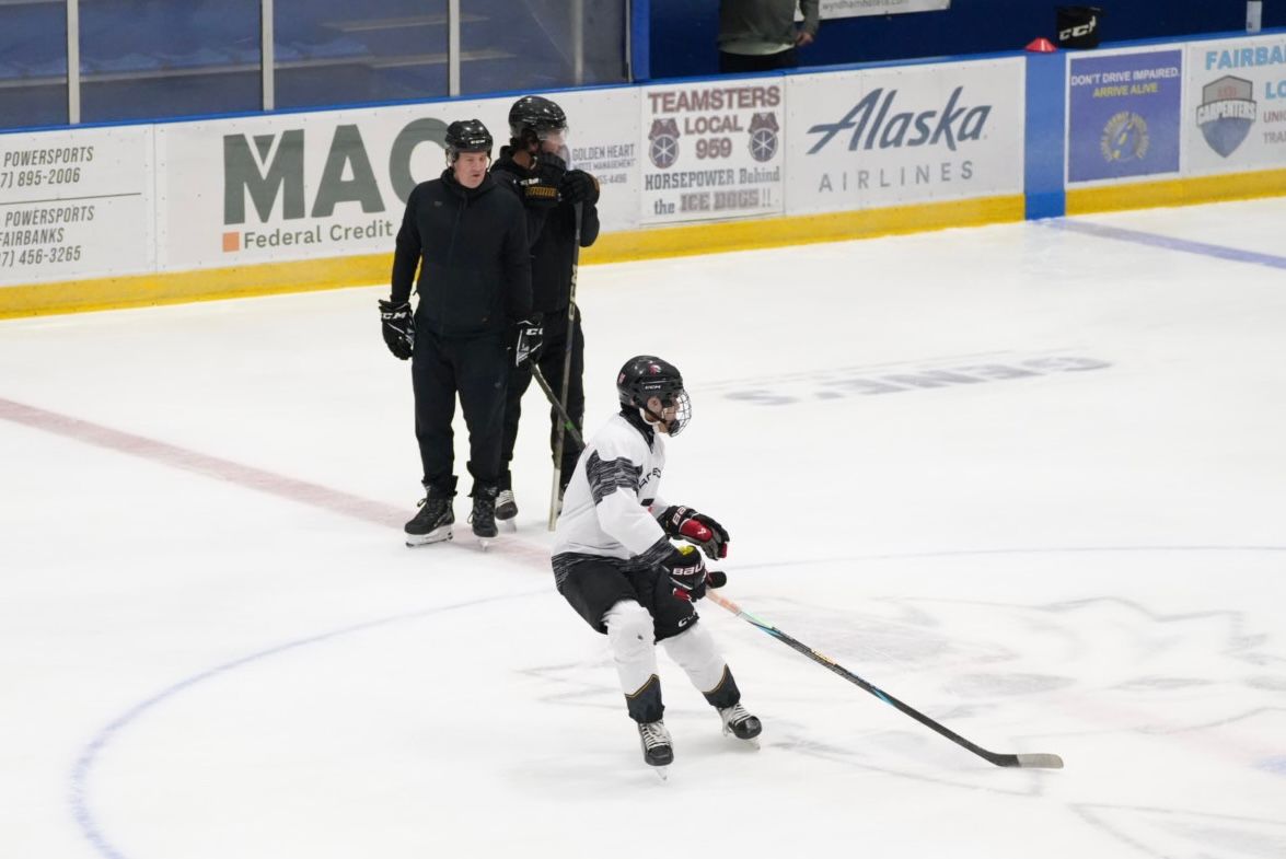 Young hockey player in white skating on ice with two coaches nearby by the boards