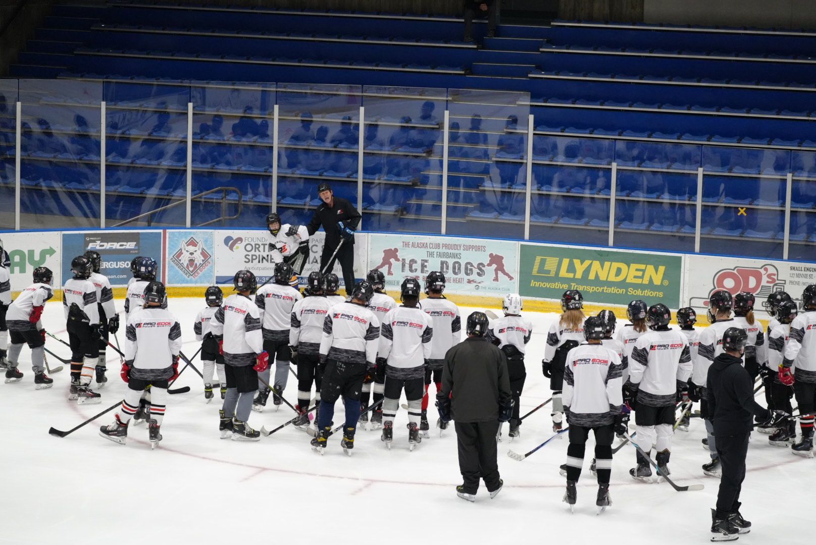 Youth hockey team gathered on the ice near the boards in an indoor arena, facing a coach.