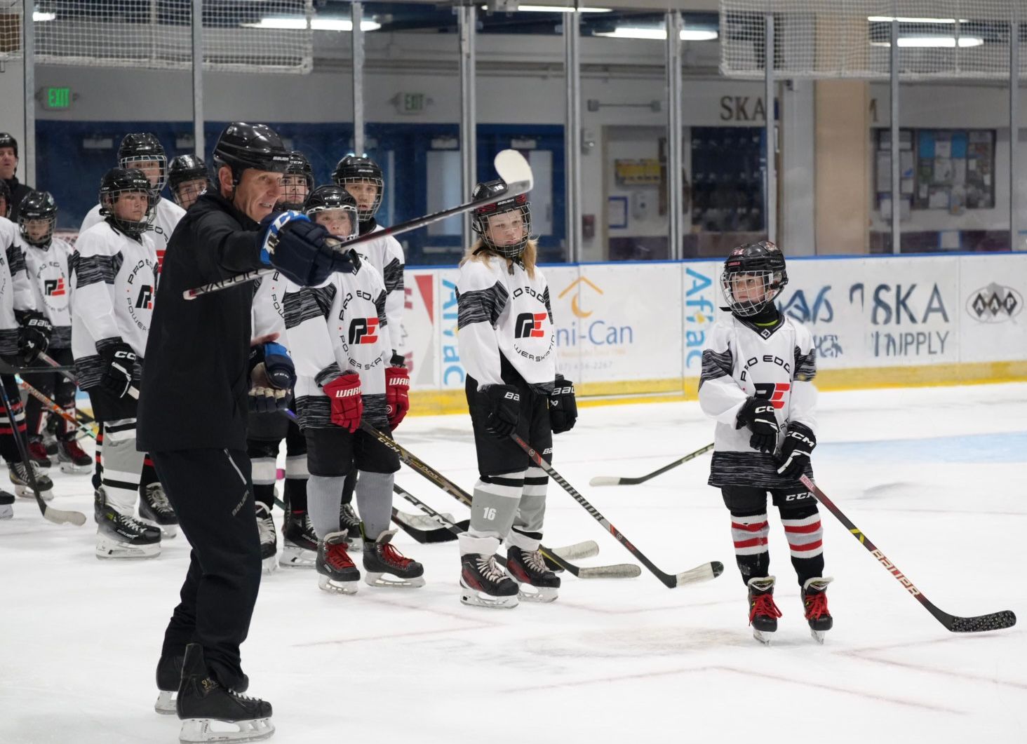 Youth hockey players and a coach on an indoor ice rink during practice, with skates and sticks visible.