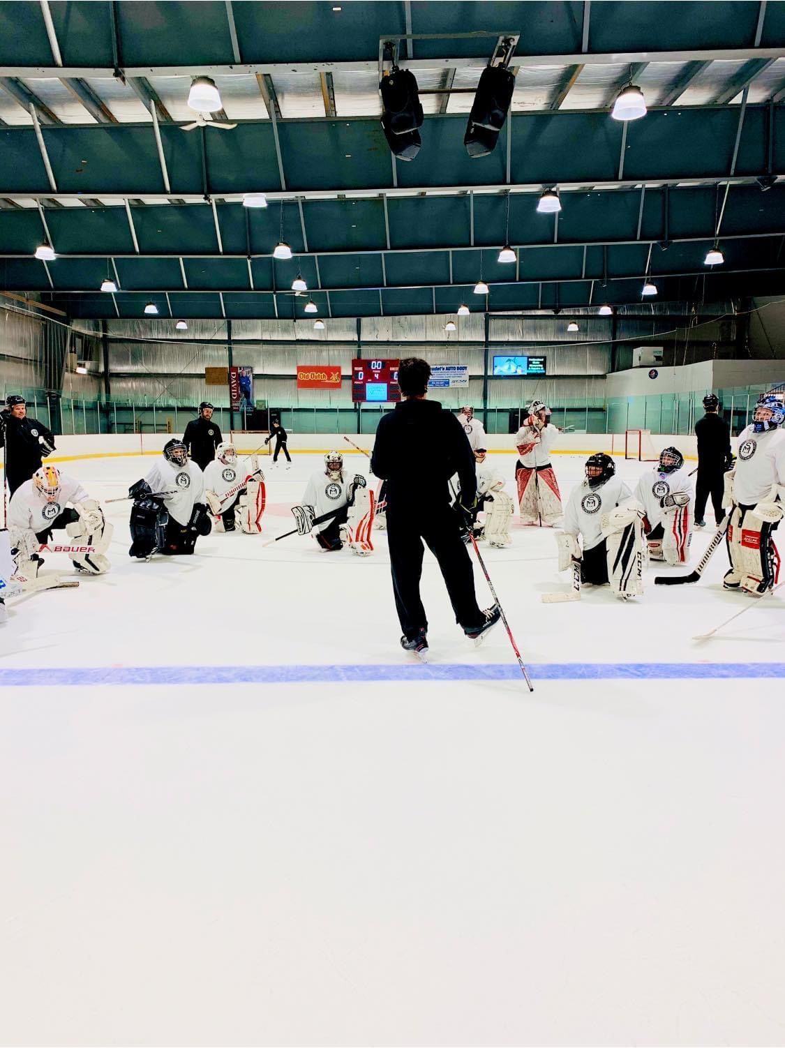 Ice hockey practice with players in white gear gathered on the rink and a coach standing in front.