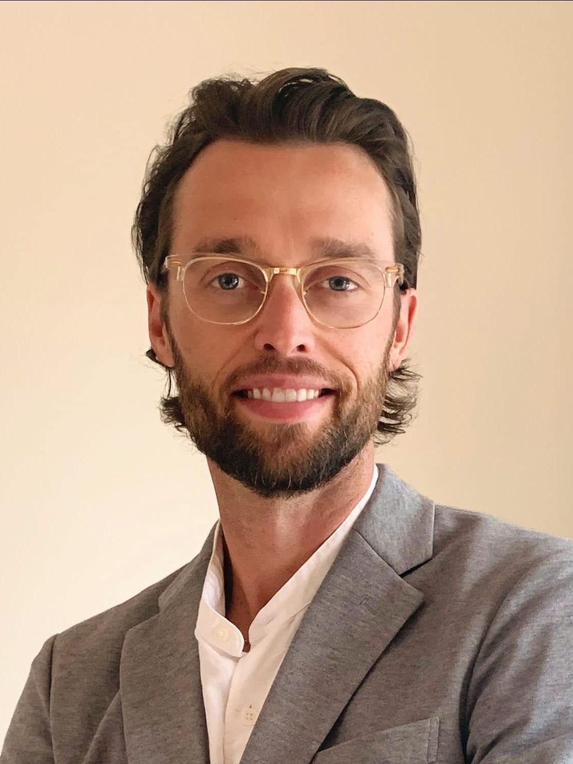 Portrait of a smiling person with glasses and a beard, wearing a gray blazer against a beige background