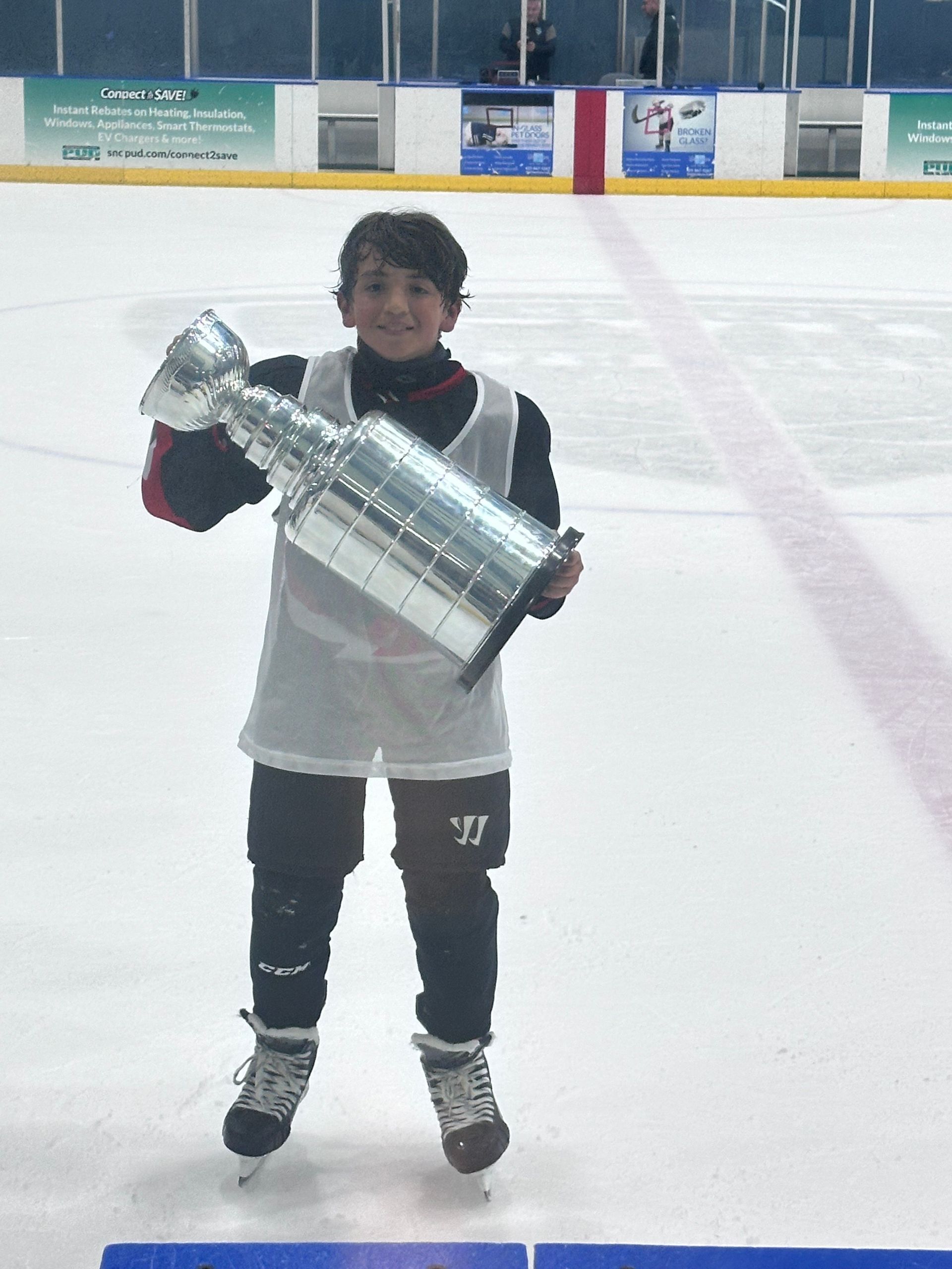 Person in hockey gear holding a large trophy on an ice rink.