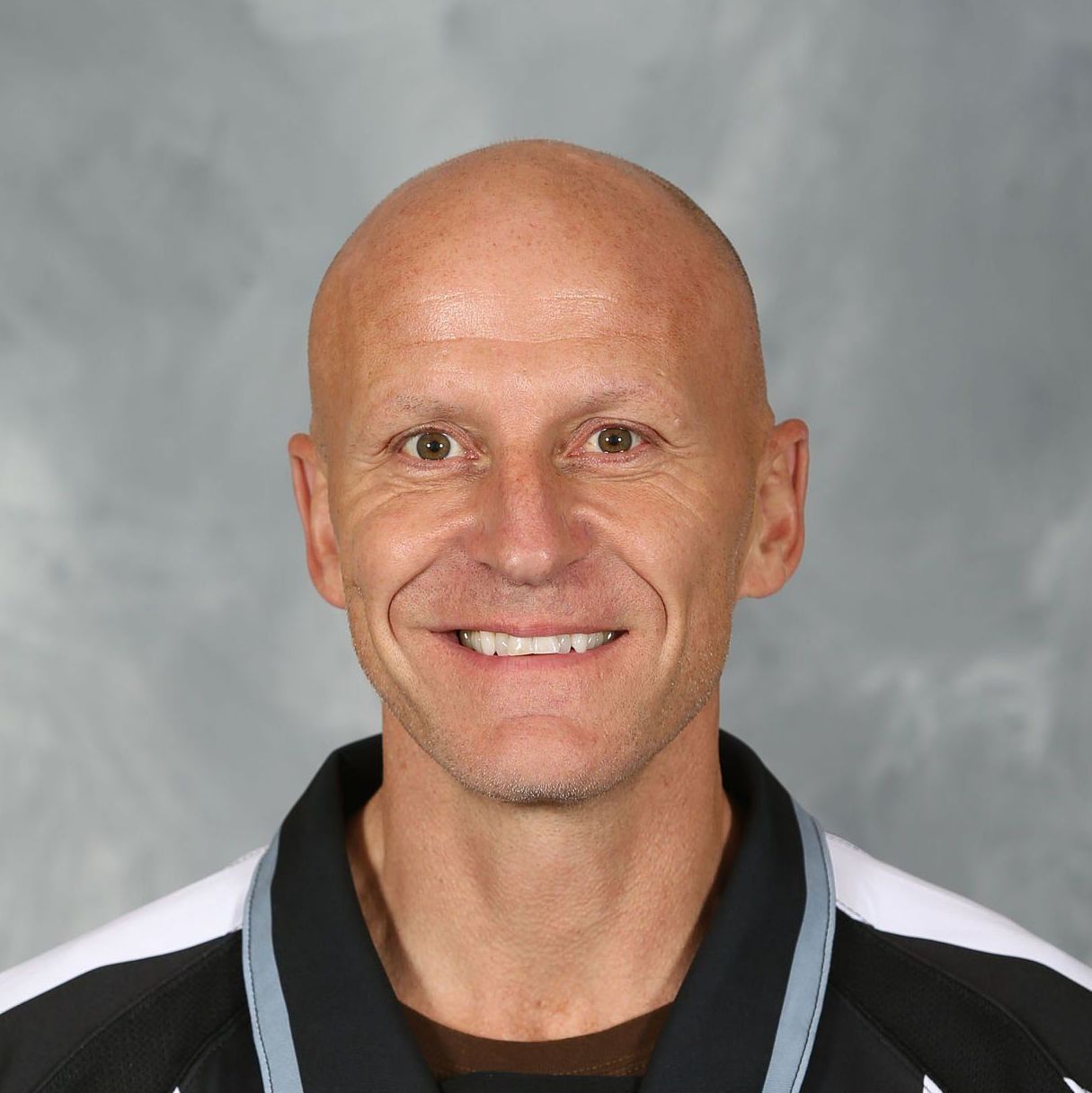 Portrait of a bald man smiling in a black and white jacket against a gray backdrop