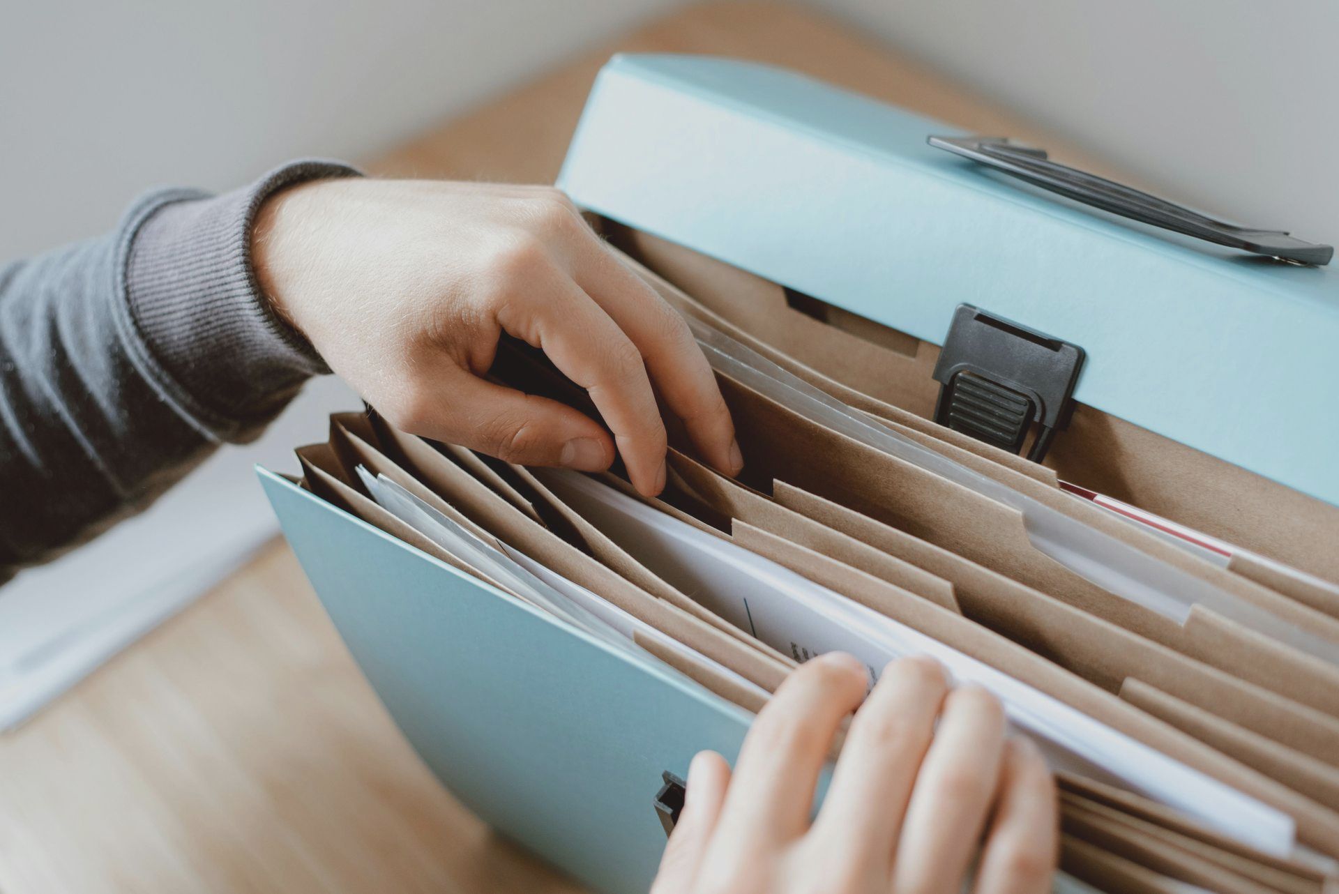Hands sorting through papers in a light blue file organizer on a wooden table.