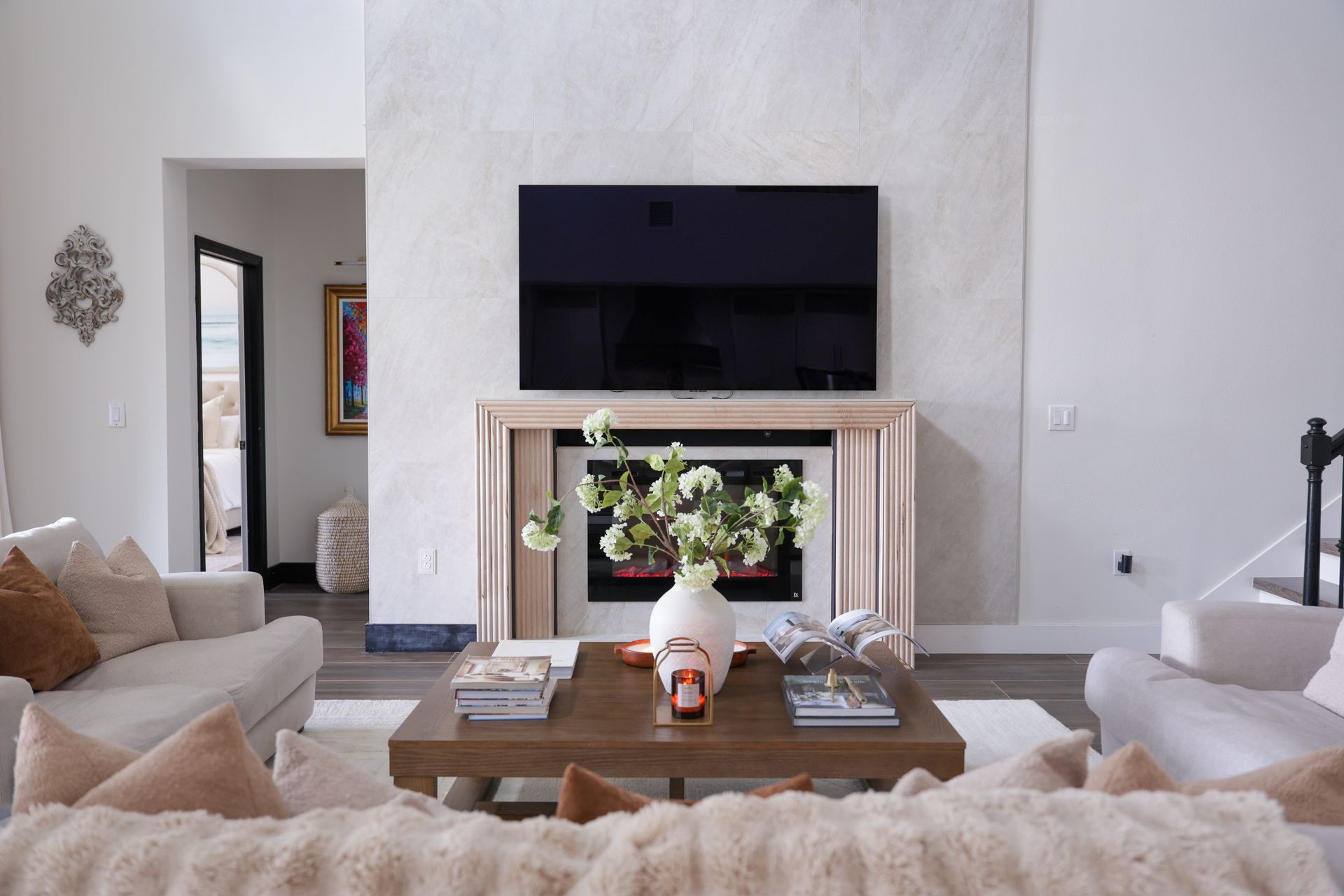 Cozy living room with white walls, fireplace, wall-mounted TV, beige sofas, and a wooden coffee table with decor.