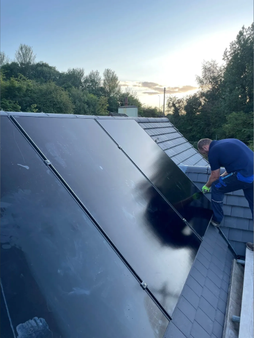 Person installing a solar panel on a gray-tiled roof, set against a backdrop of trees and a cloudy sky.