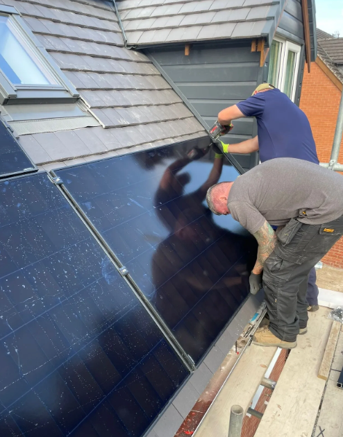 Two workers installing solar panels on a slate roof. One cuts, the other works near the panels.