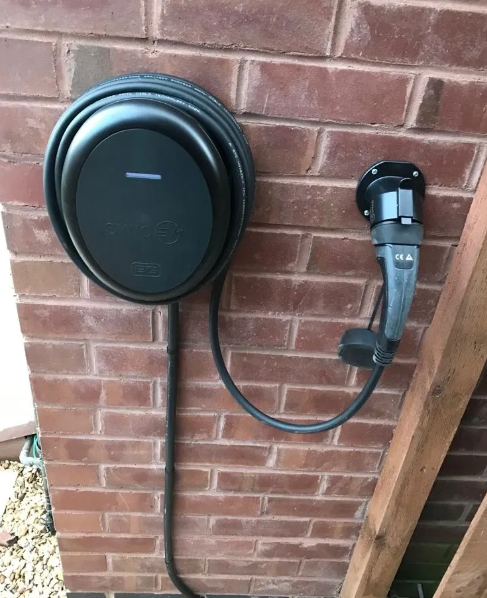 Black EV charger mounted on a brick wall with cable wrapped around the unit and a plug attached.