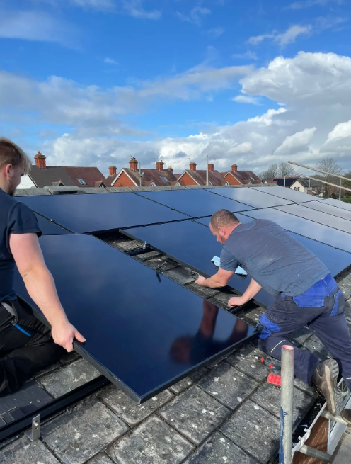 Two workers installing solar panels on a slate rooftop under a partly cloudy sky.