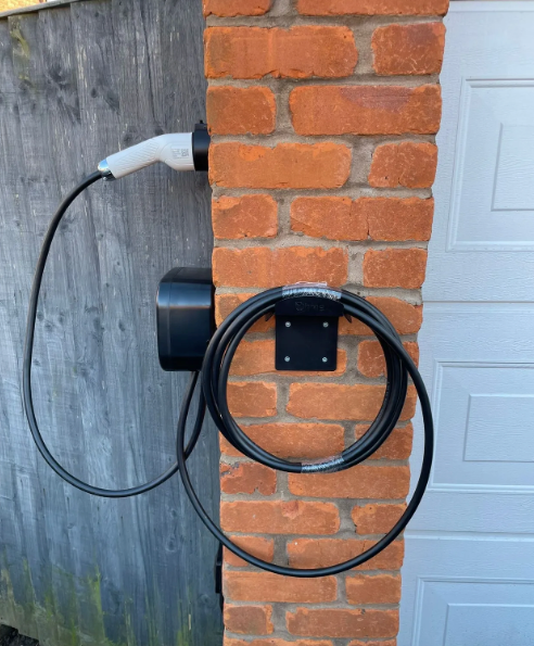 An electric vehicle charger mounted on a brick pillar next to a wooden fence and a white garage door.