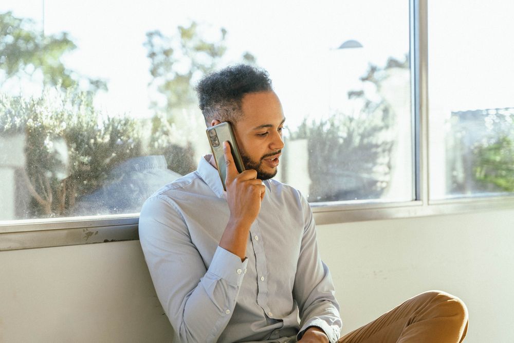 Man in light blue shirt talking on a cell phone near a window.