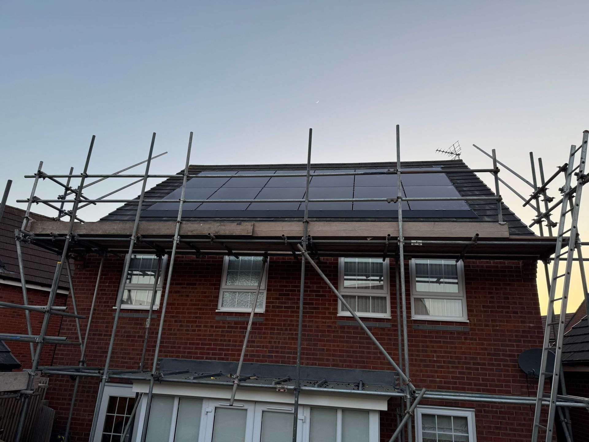 Solar panels installed on a red brick house roof with scaffolding. Evening sky.