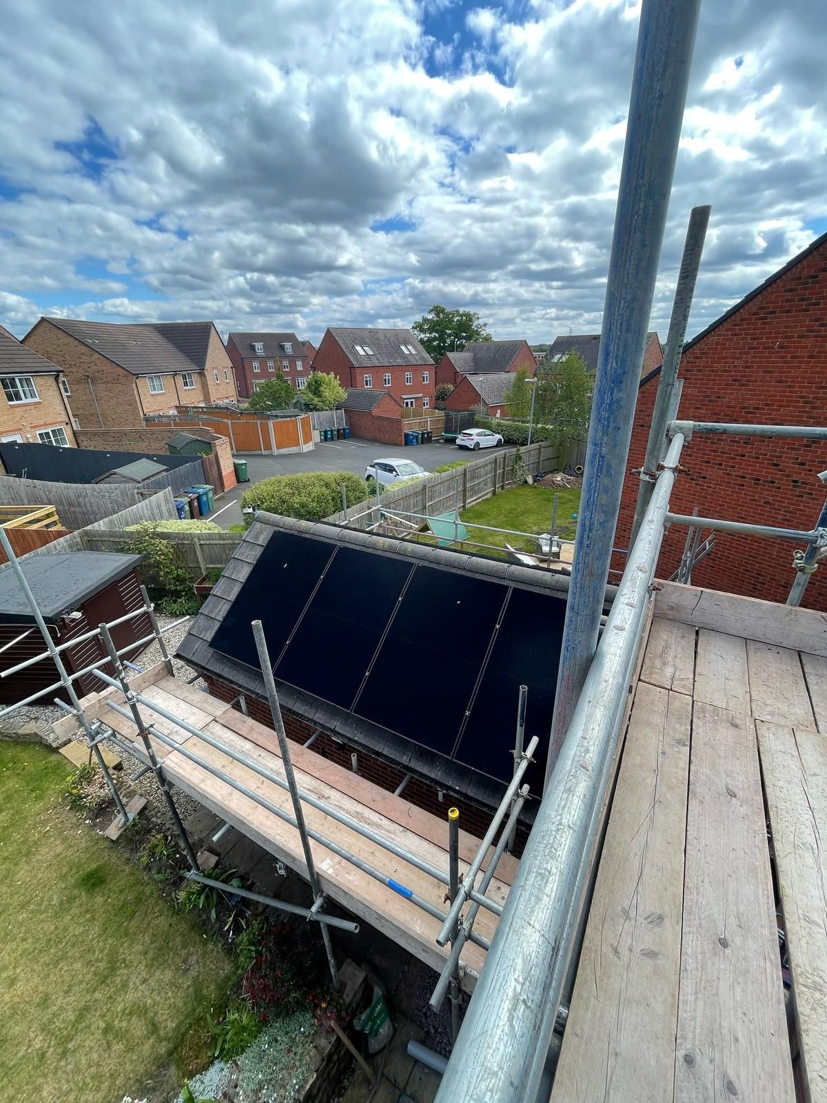 View from scaffolding of solar panels on a house roof. Backyard with houses, grass, and sky with clouds.