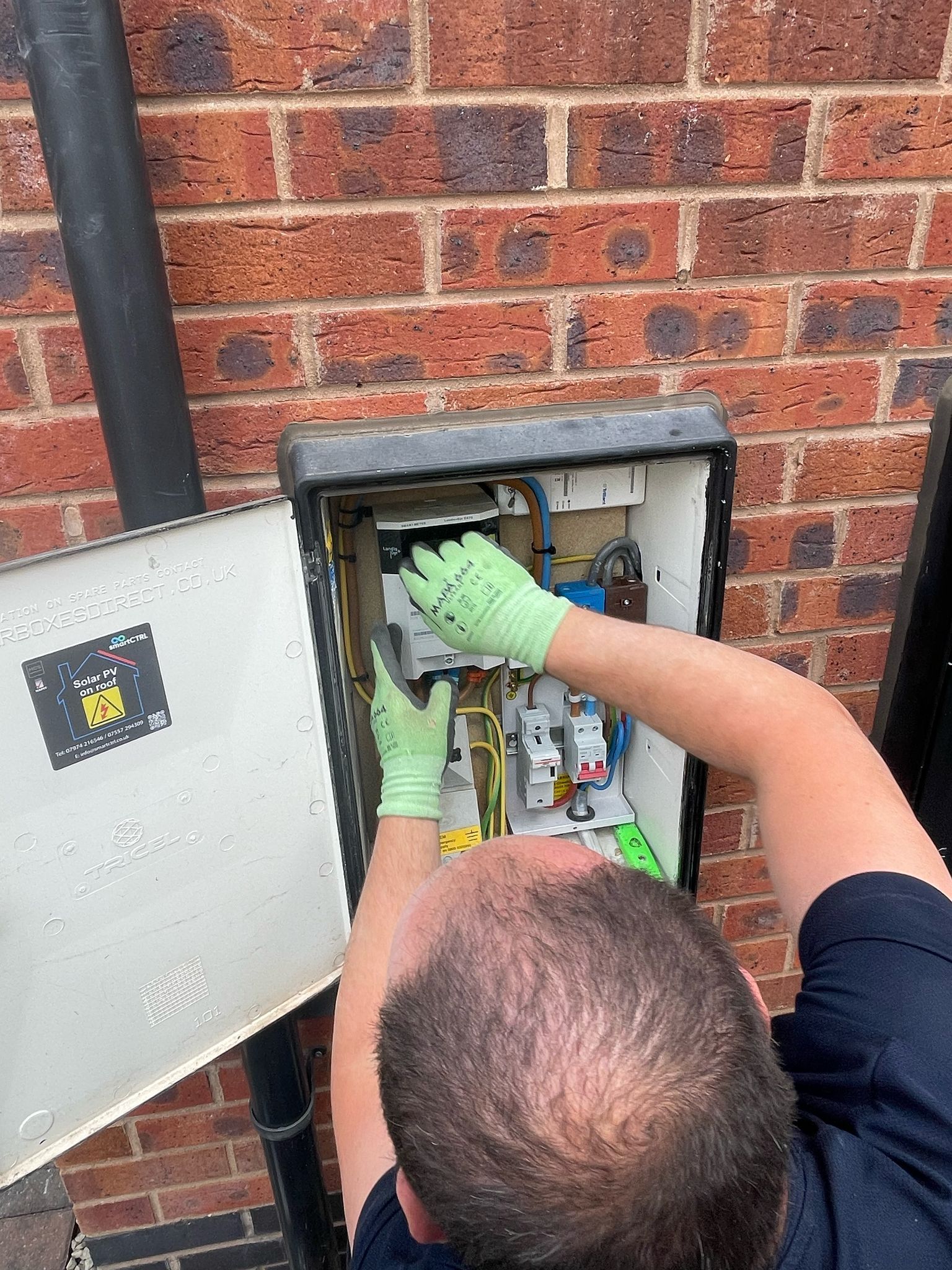 Electrician working on a gray electrical box mounted on a brick wall. He wears gloves and opens the panel.
