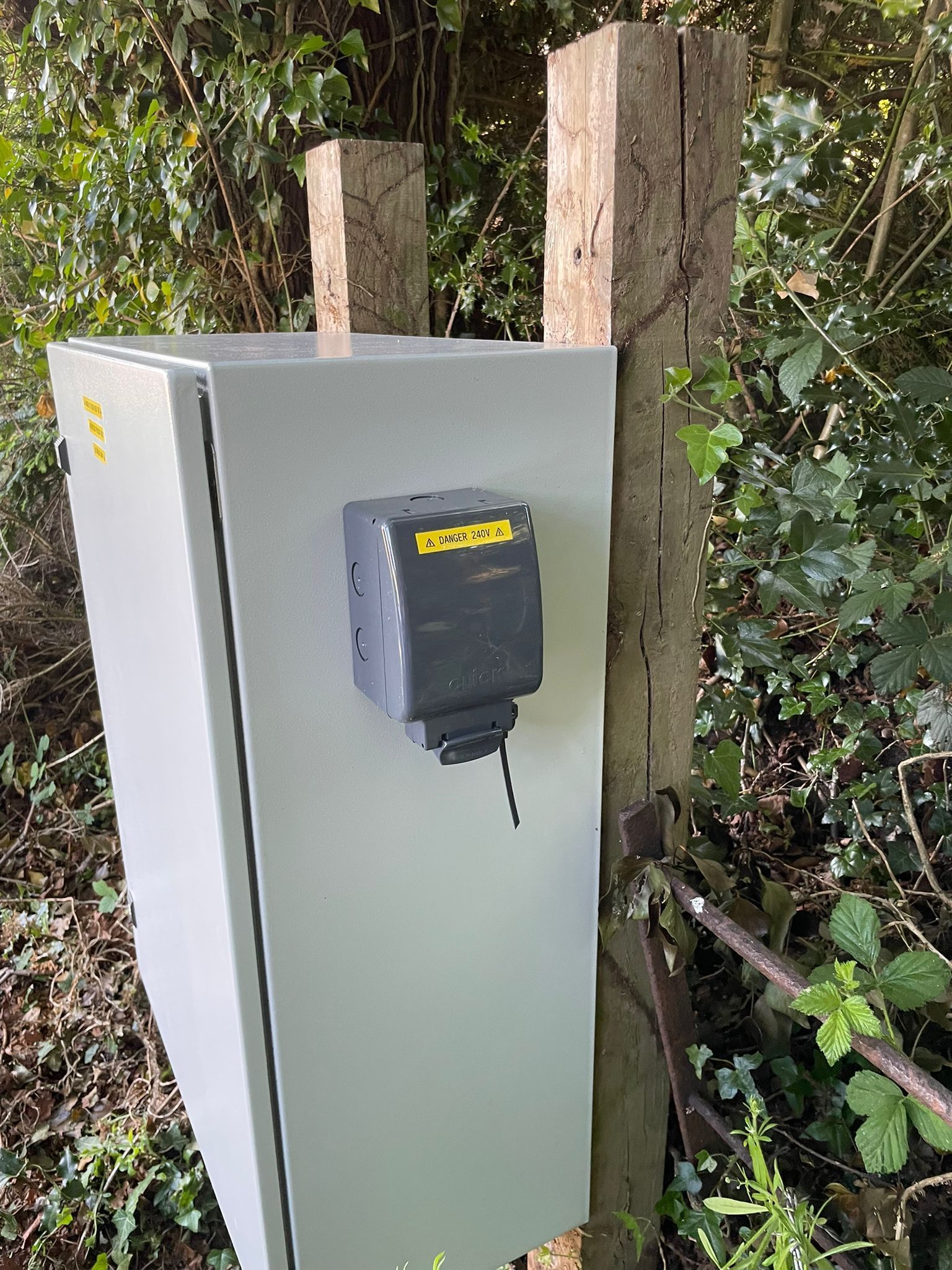 Gray electrical box with attached dark grey device mounted on wooden posts in a wooded area.