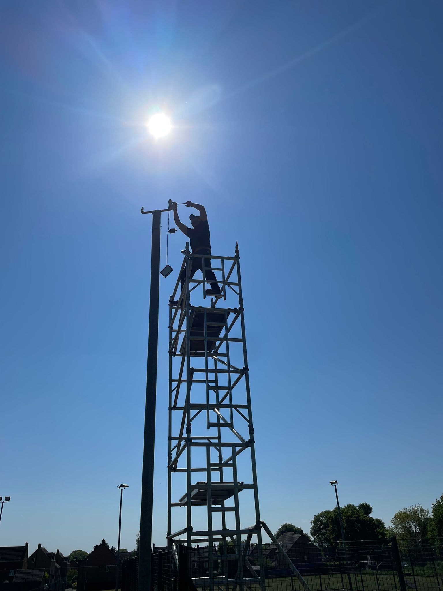 Person on scaffolding, installing fixture on tall pole, under bright sun.