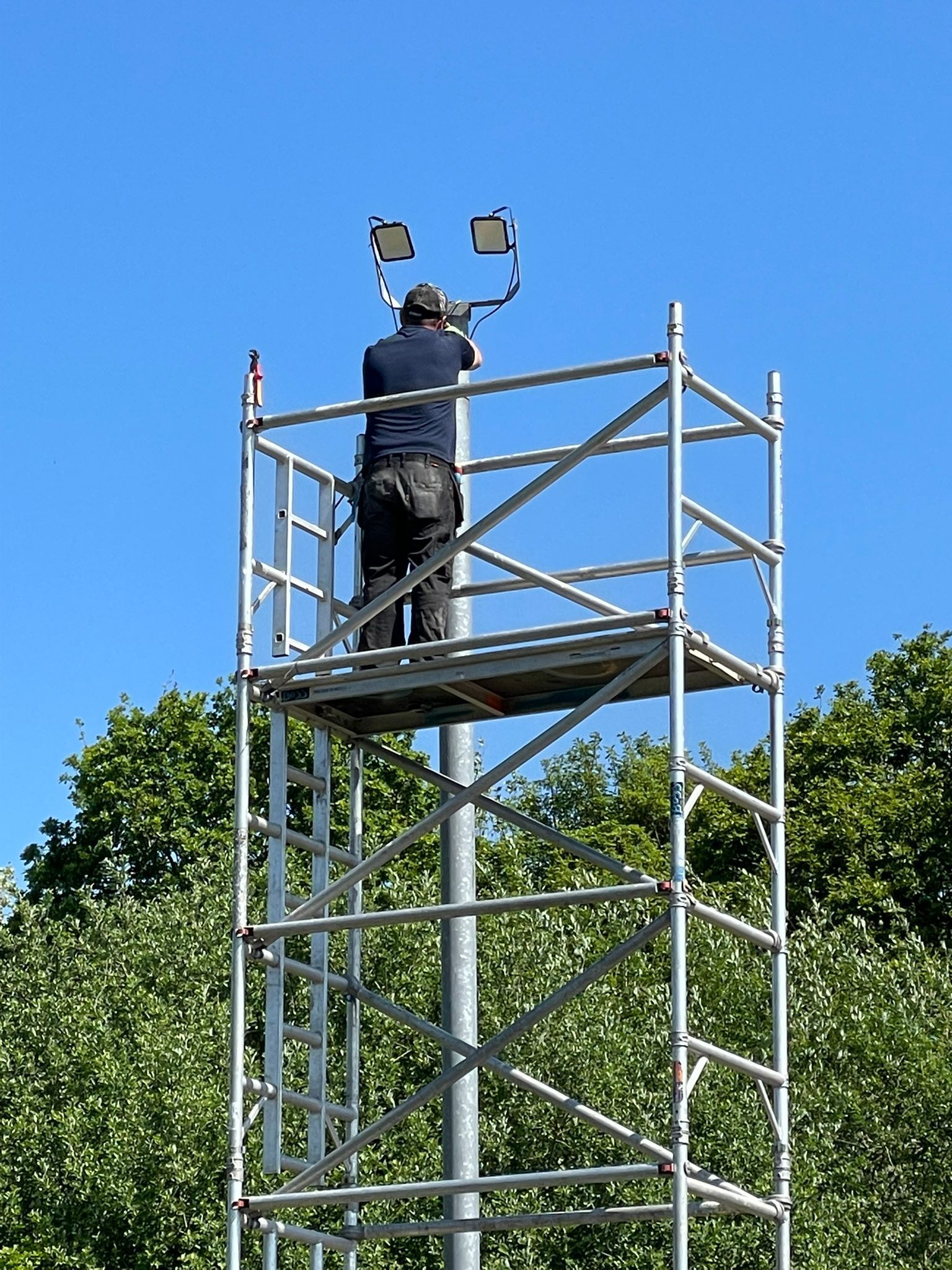 Man on scaffolding installing equipment on a tall metal pole. Blue sky, green trees in background.