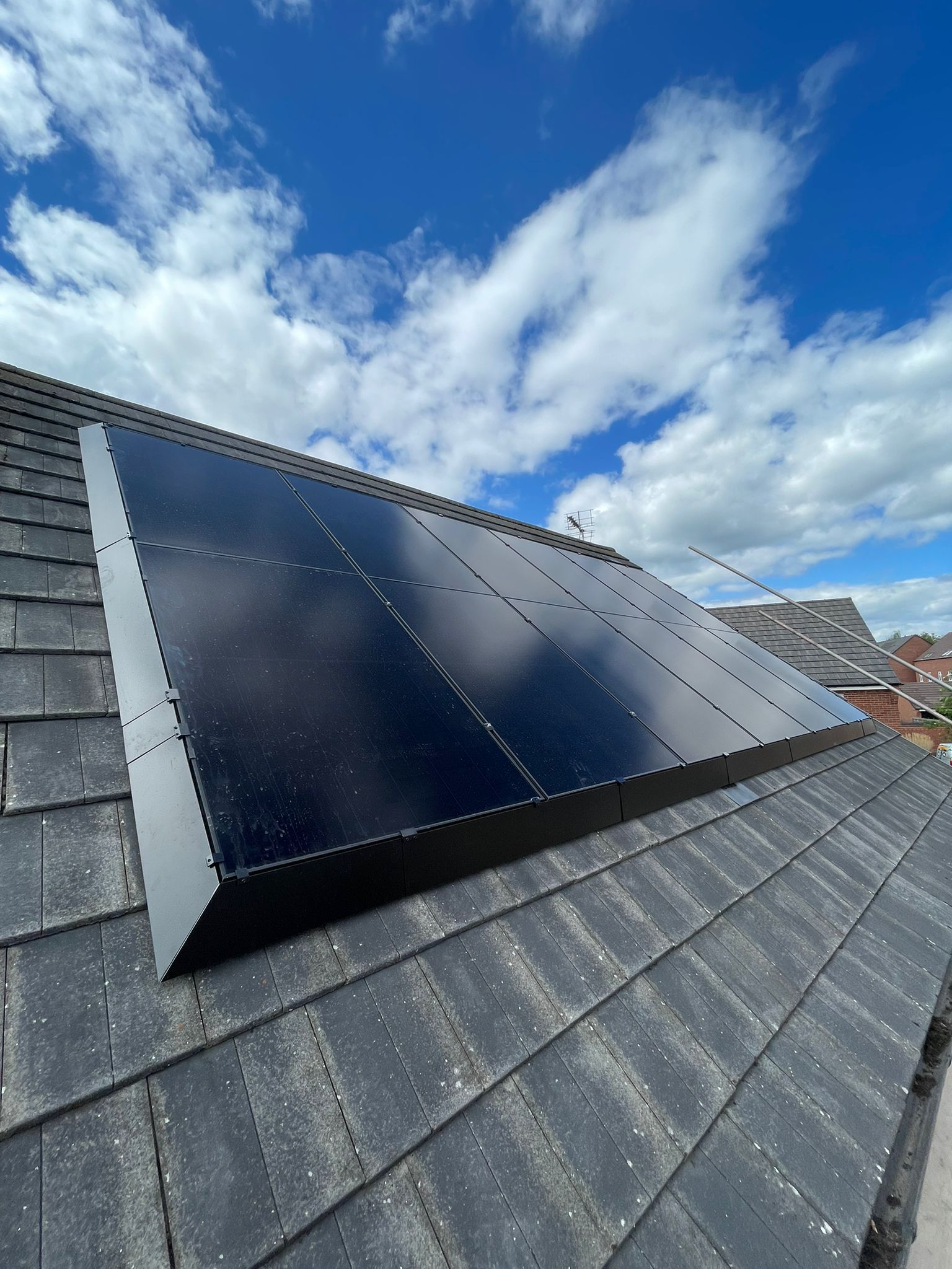 Solar panels installed on a dark grey tiled roof, against a cloudy blue sky.
