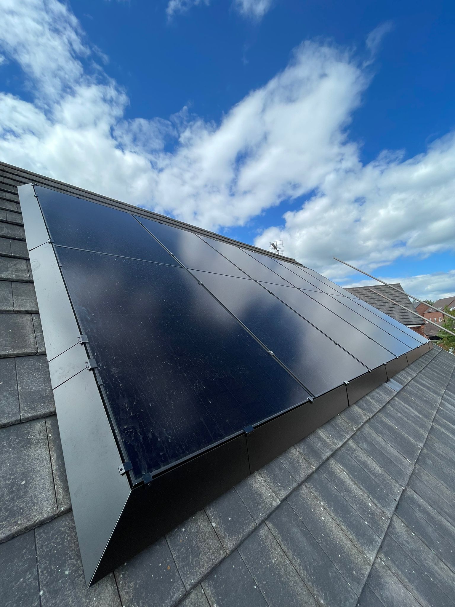 Solar panels installed on a dark grey tiled roof, against a cloudy blue sky.