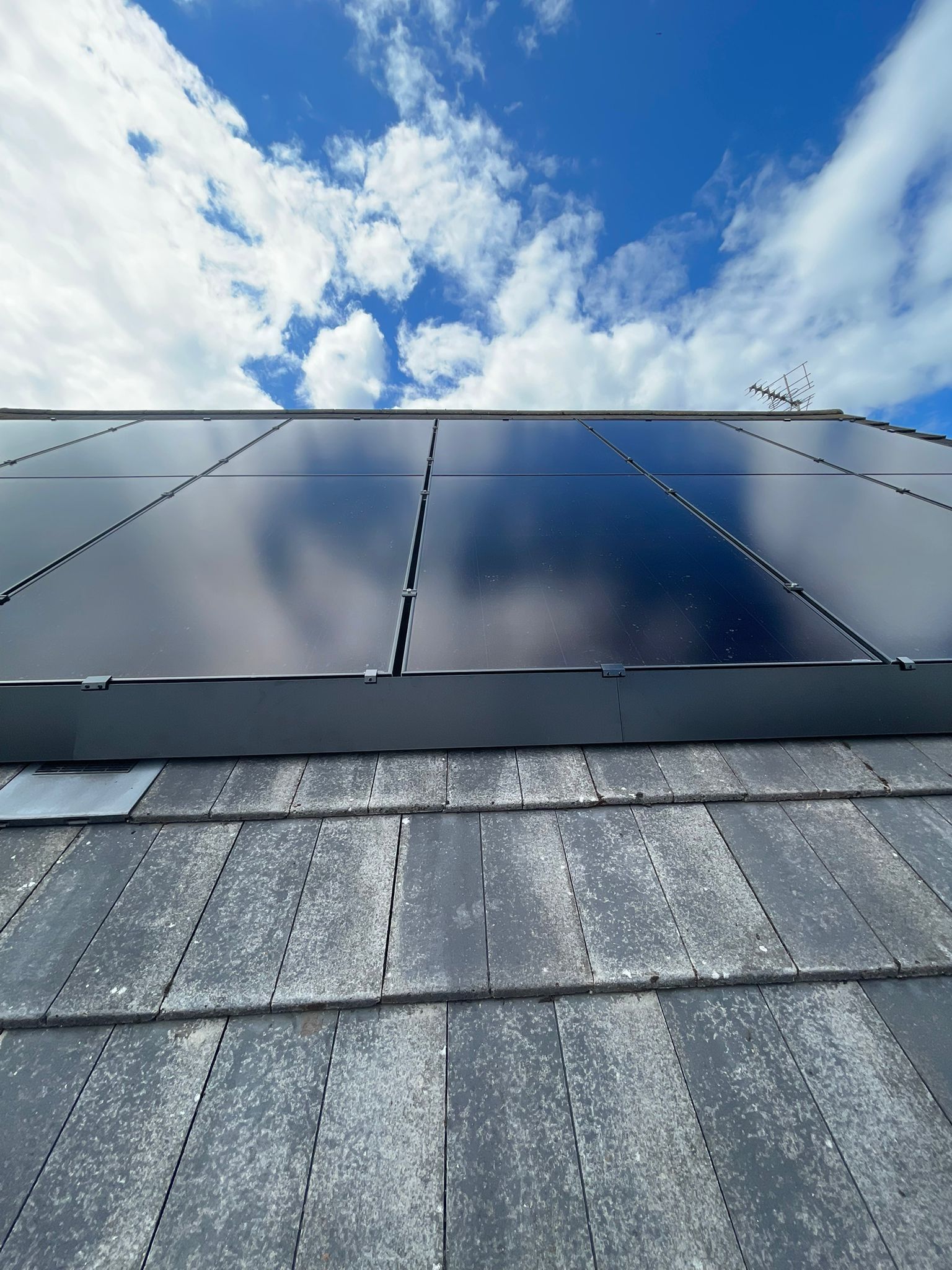 Solar panels installed on a dark grey tiled roof against a bright blue sky with clouds.