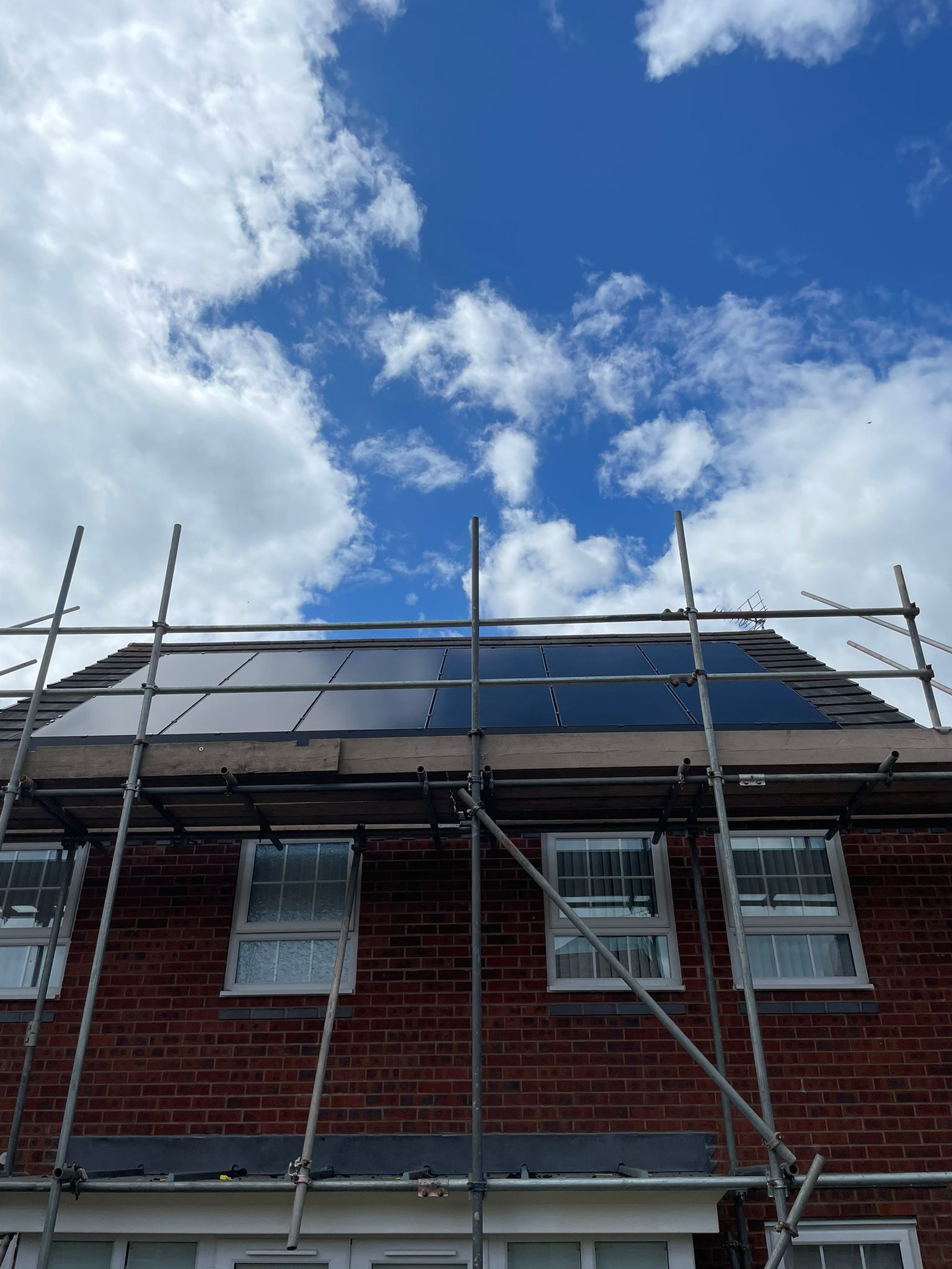 Solar panels installed on a brick house roof under a cloudy blue sky, scaffolding surrounding the roof.