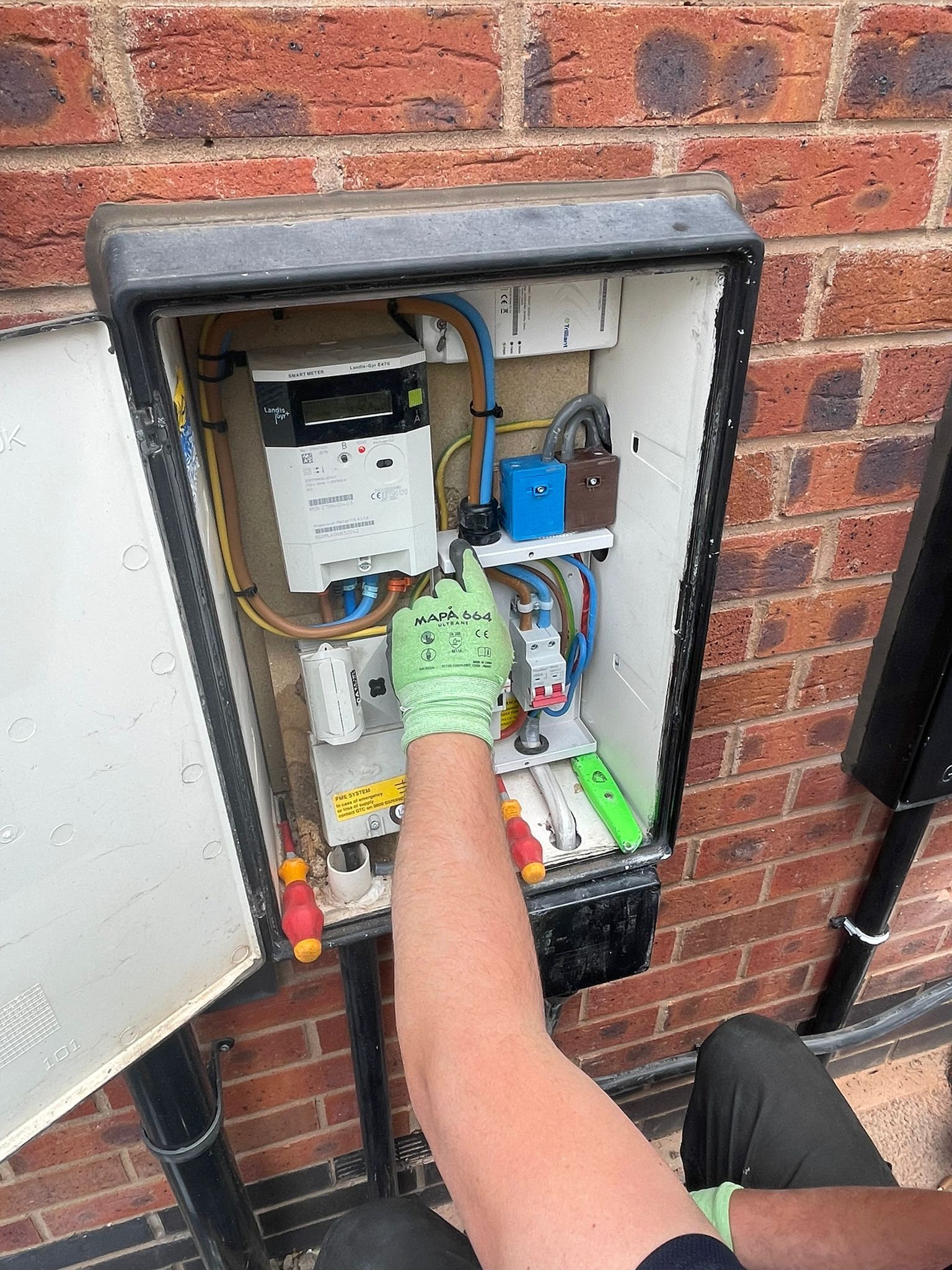Person in green gloves reaching into an open electrical box on a brick wall.