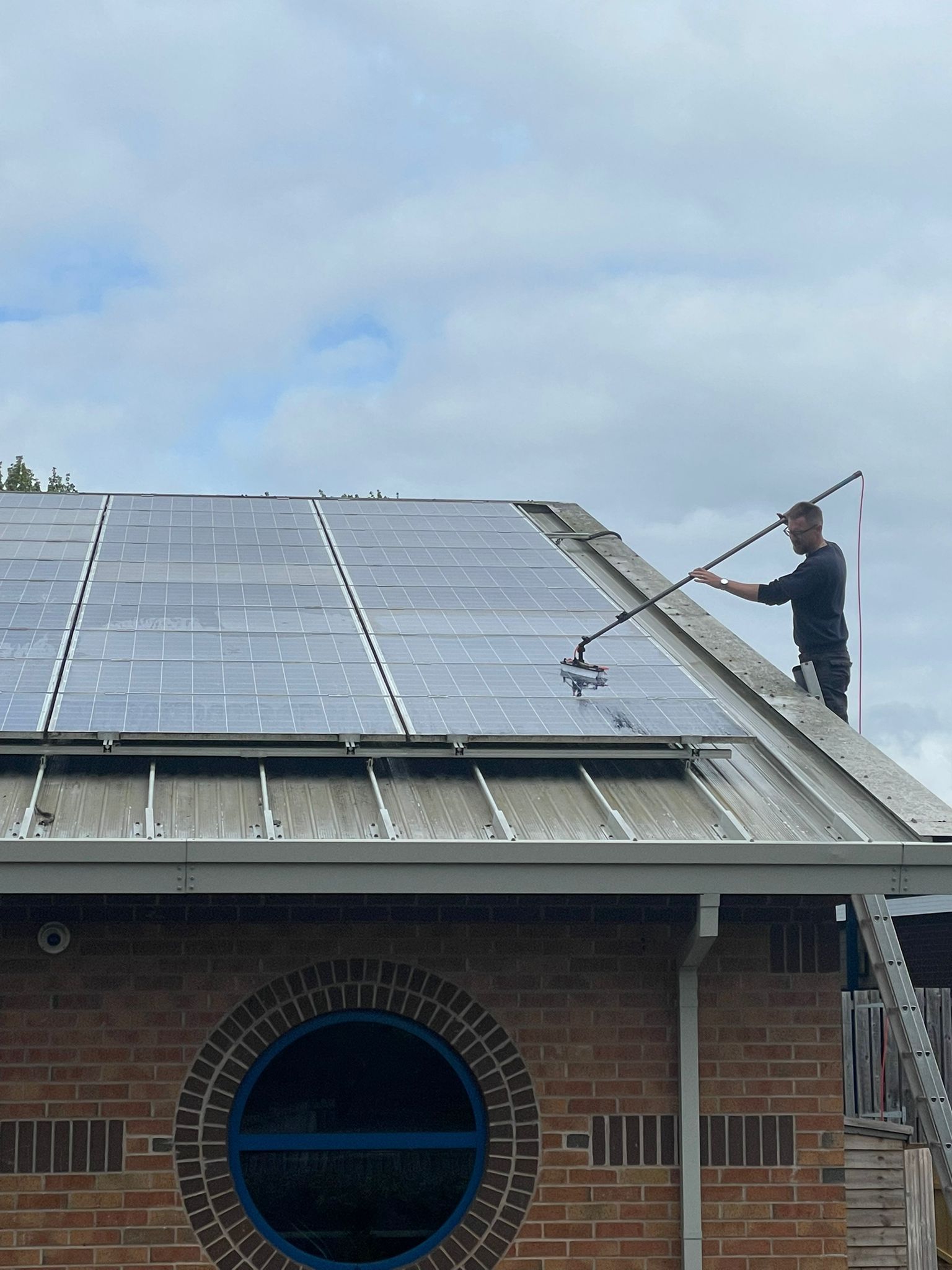 Man cleaning solar panels on a building's roof with a brush on a long pole. Cloudy sky.