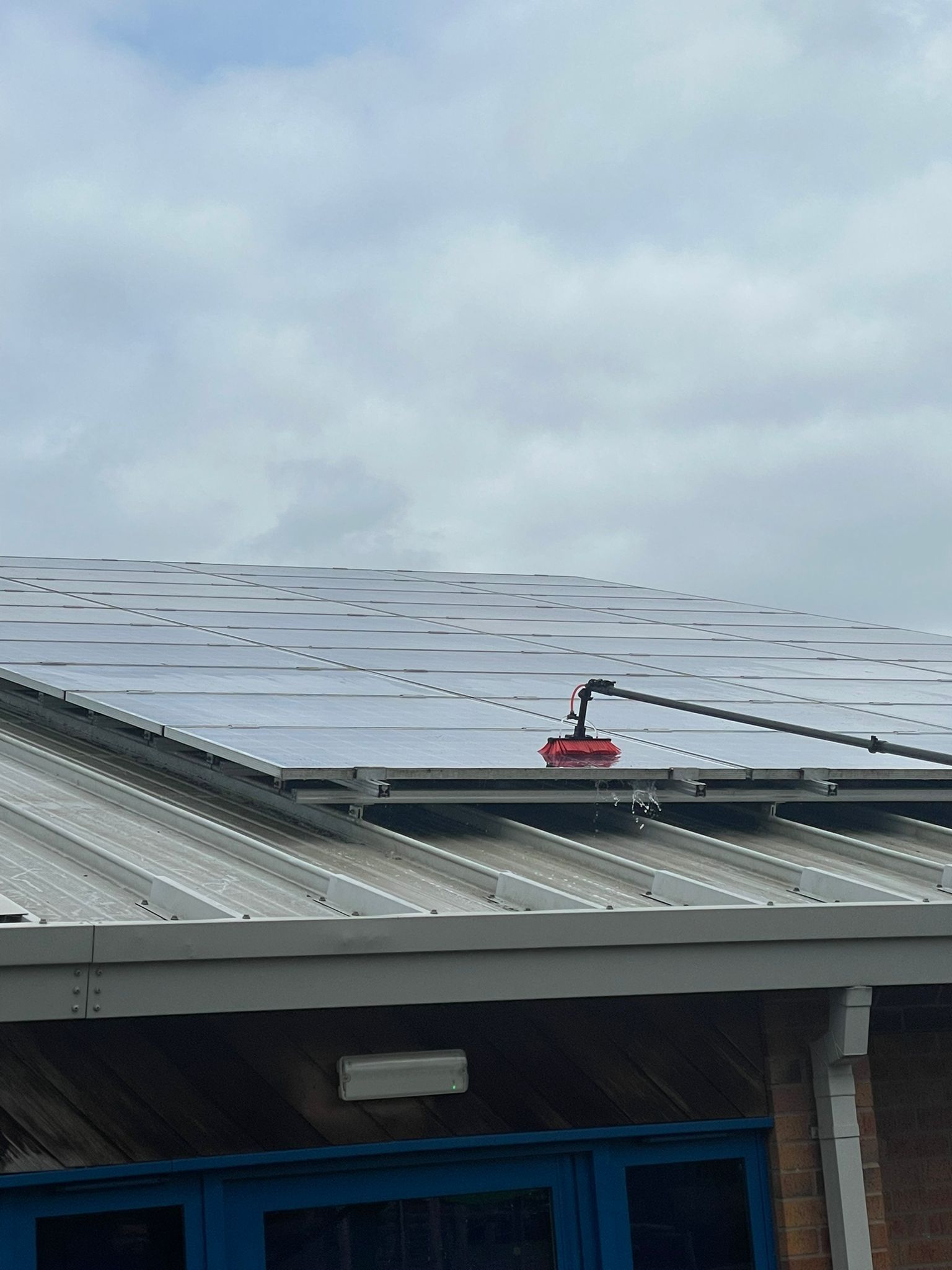 A red robot cleaning solar panels on a gray industrial building roof under a cloudy sky.