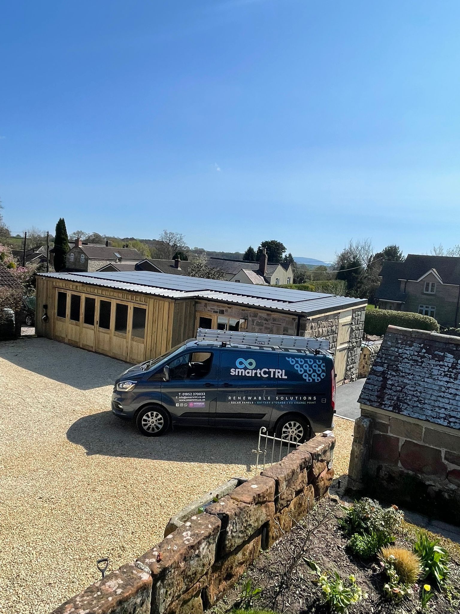 Blue van parked on gravel driveway. Wooden structure with solar panels in background, houses, blue sky.