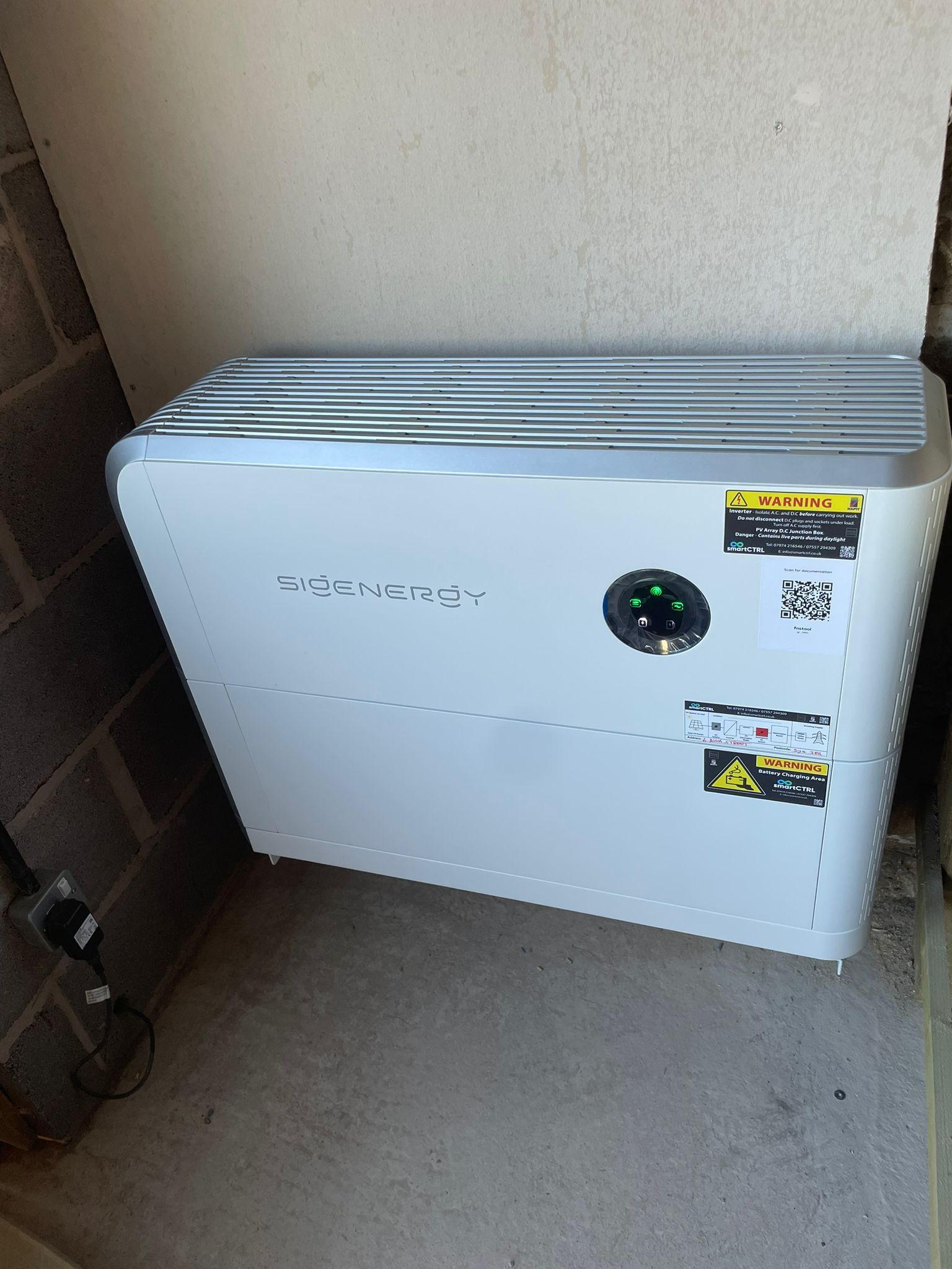 White solar panel inverter mounted on a brick wall. Black circular indicator light on the front.