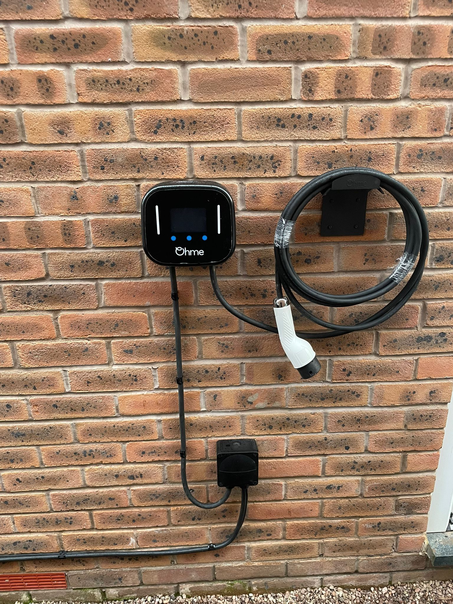 Black EV charger mounted on a brick wall with cable and plug, next to a black electrical box.