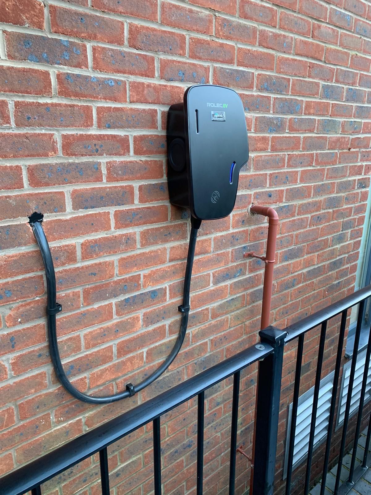 Black electric vehicle charging station mounted on a red brick wall. Cable runs down to a railing.