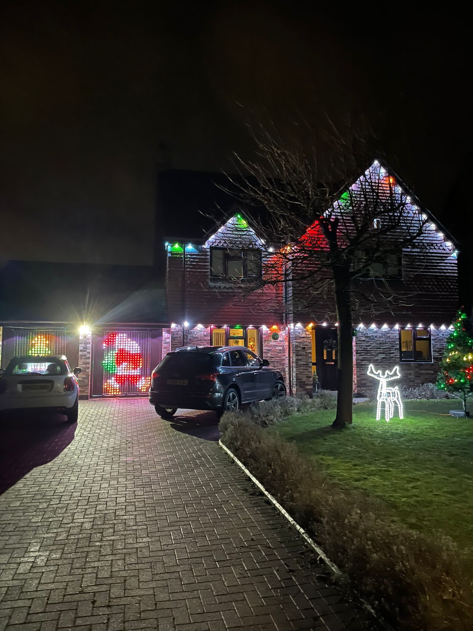 House decorated with Christmas lights at night; cars in driveway; Santa figure.