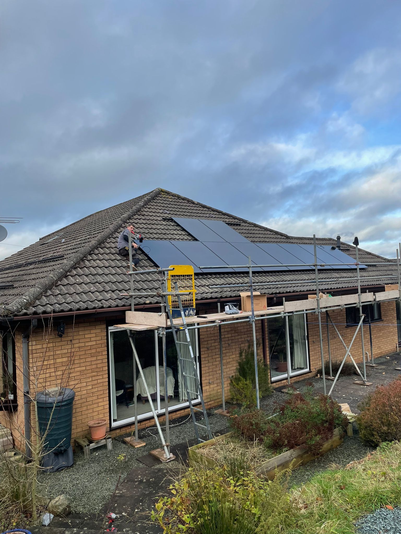 Workers installing solar panels on a residential rooftop, with scaffolding in place. Overcast sky.