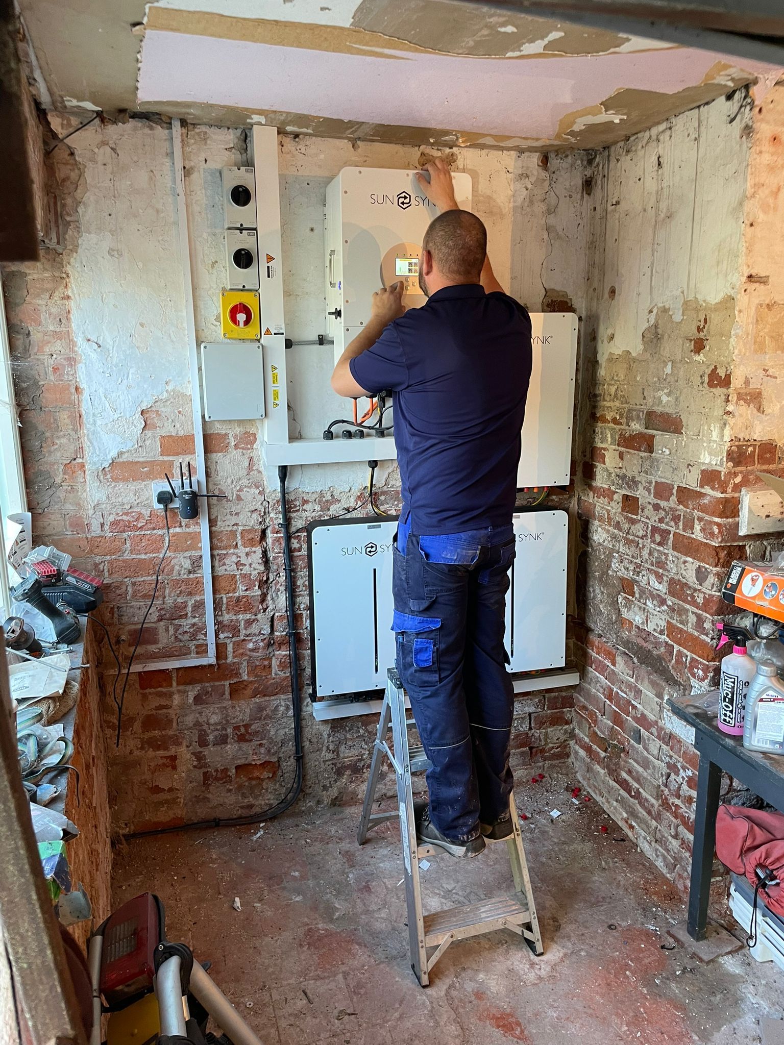 Man on ladder installing equipment in a brick-walled room. Wall paint is peeling.